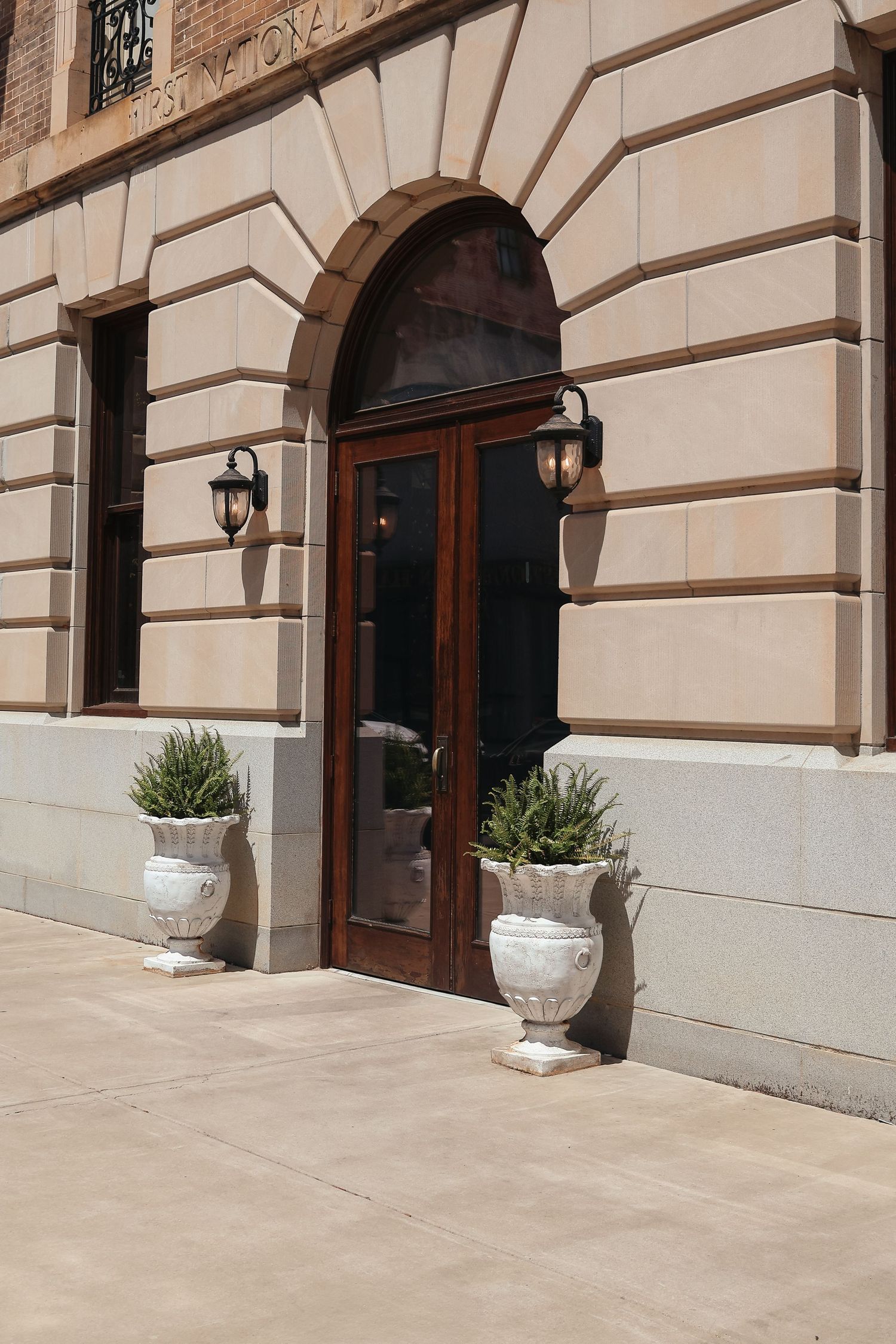 Elegant stone building entrance with arched wooden doors, ornate wall lanterns, and decorative white urns with greenery.