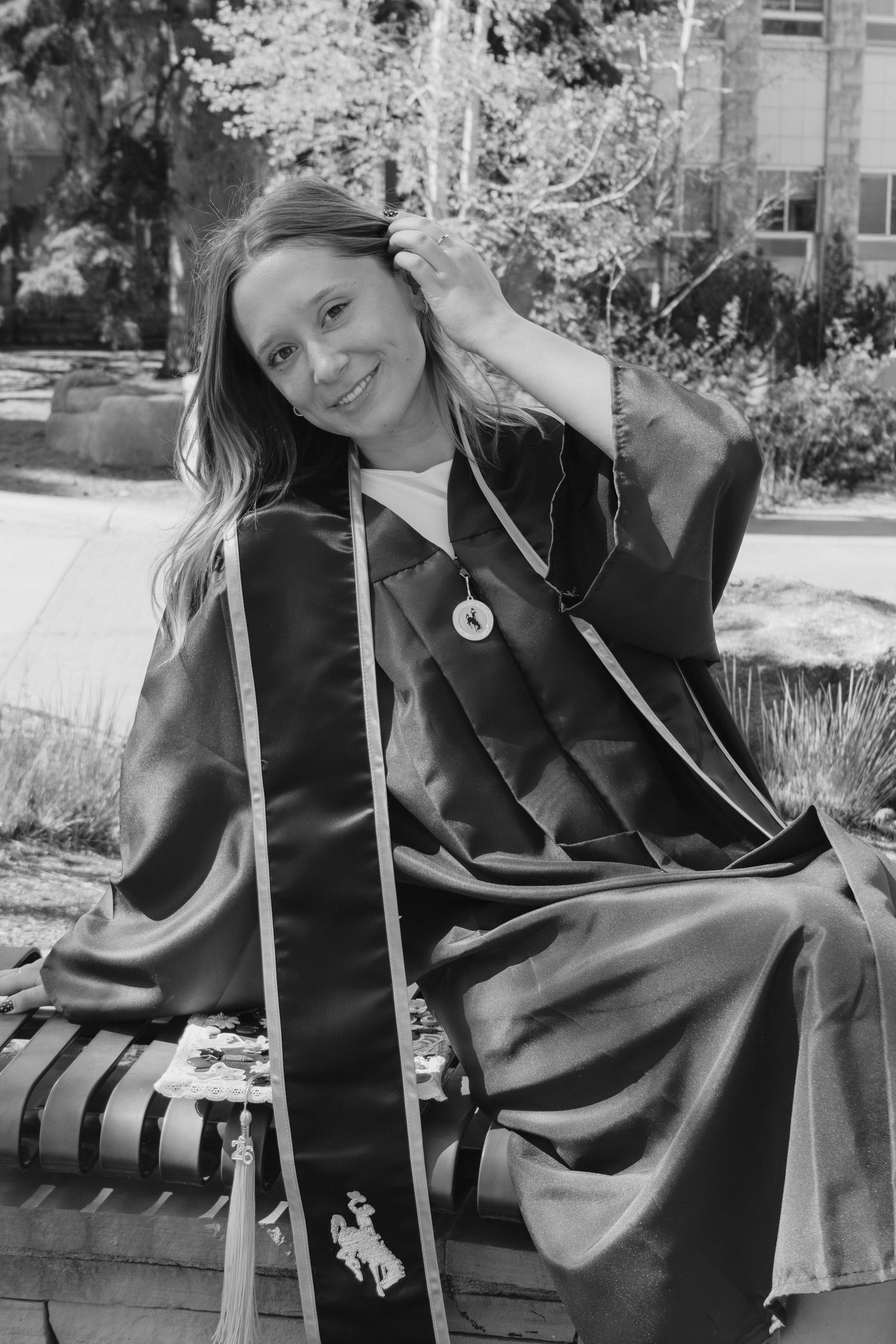 Young woman in graduation gown and stole smiles while seated outdoors in black and white photo.