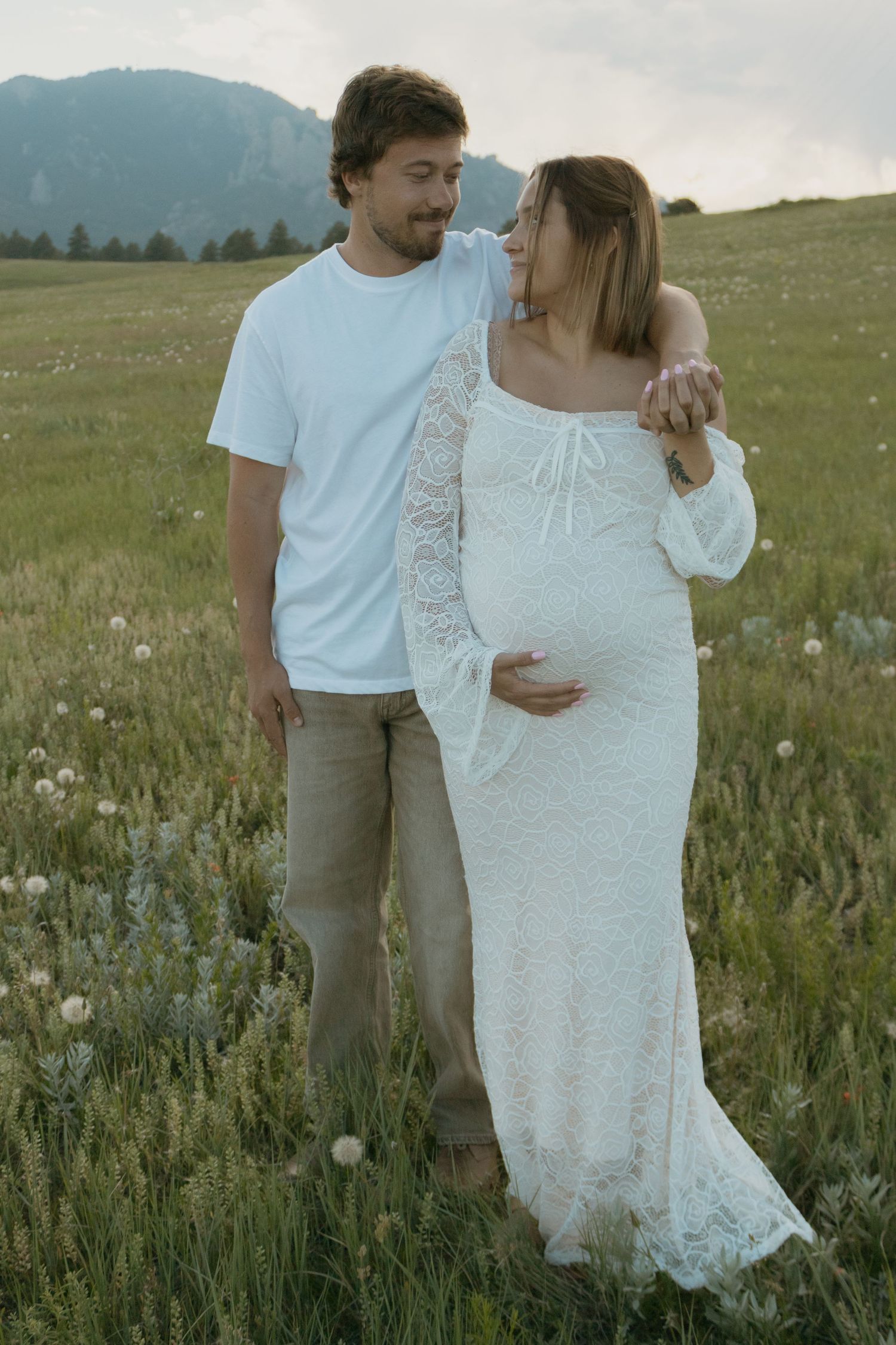 A couple poses in a green meadow, the woman in a white lace maternity gown cradling her bump, mountains in the background.