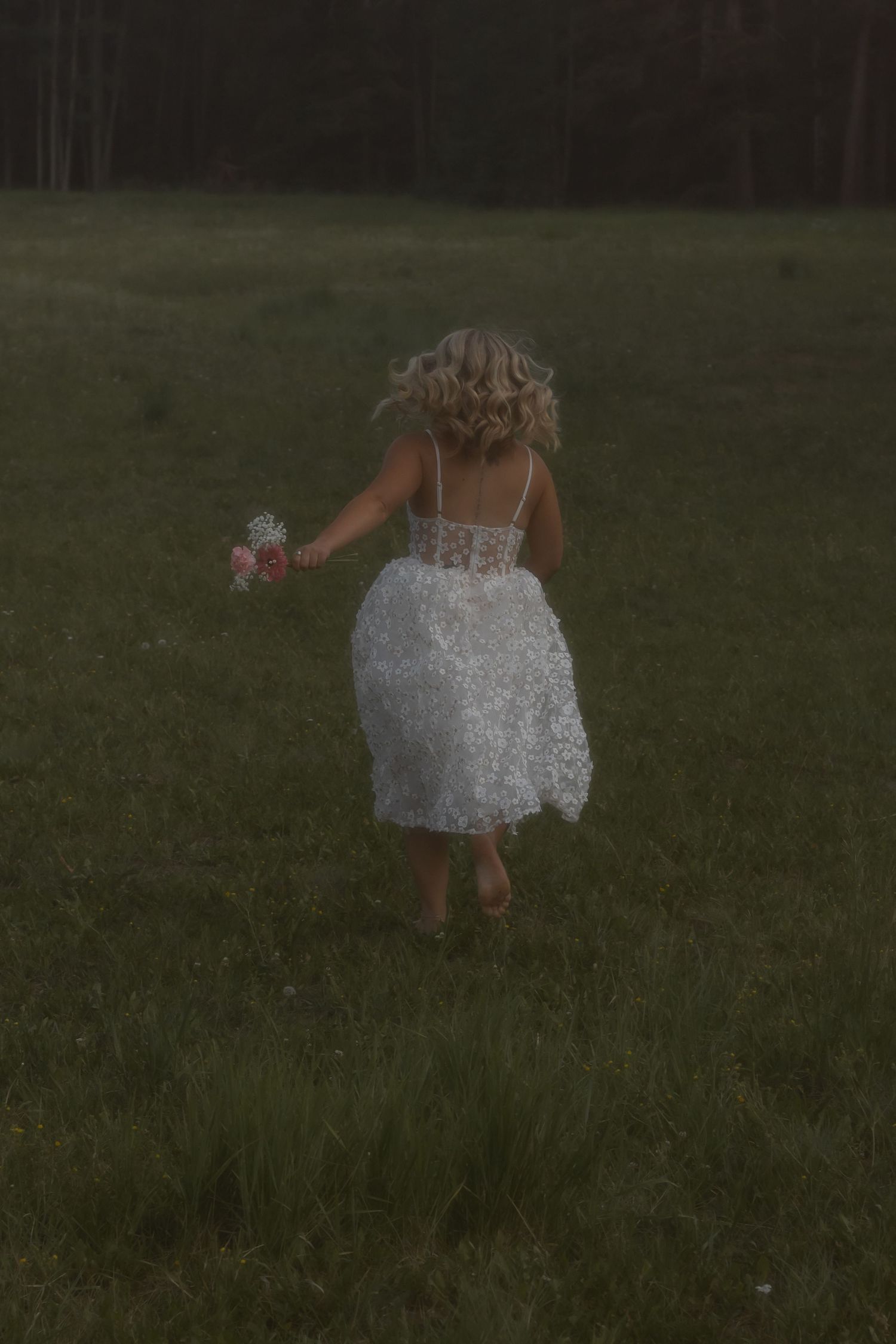 A young girl in a white lace dress runs through a green meadow holding flowers at dusk.