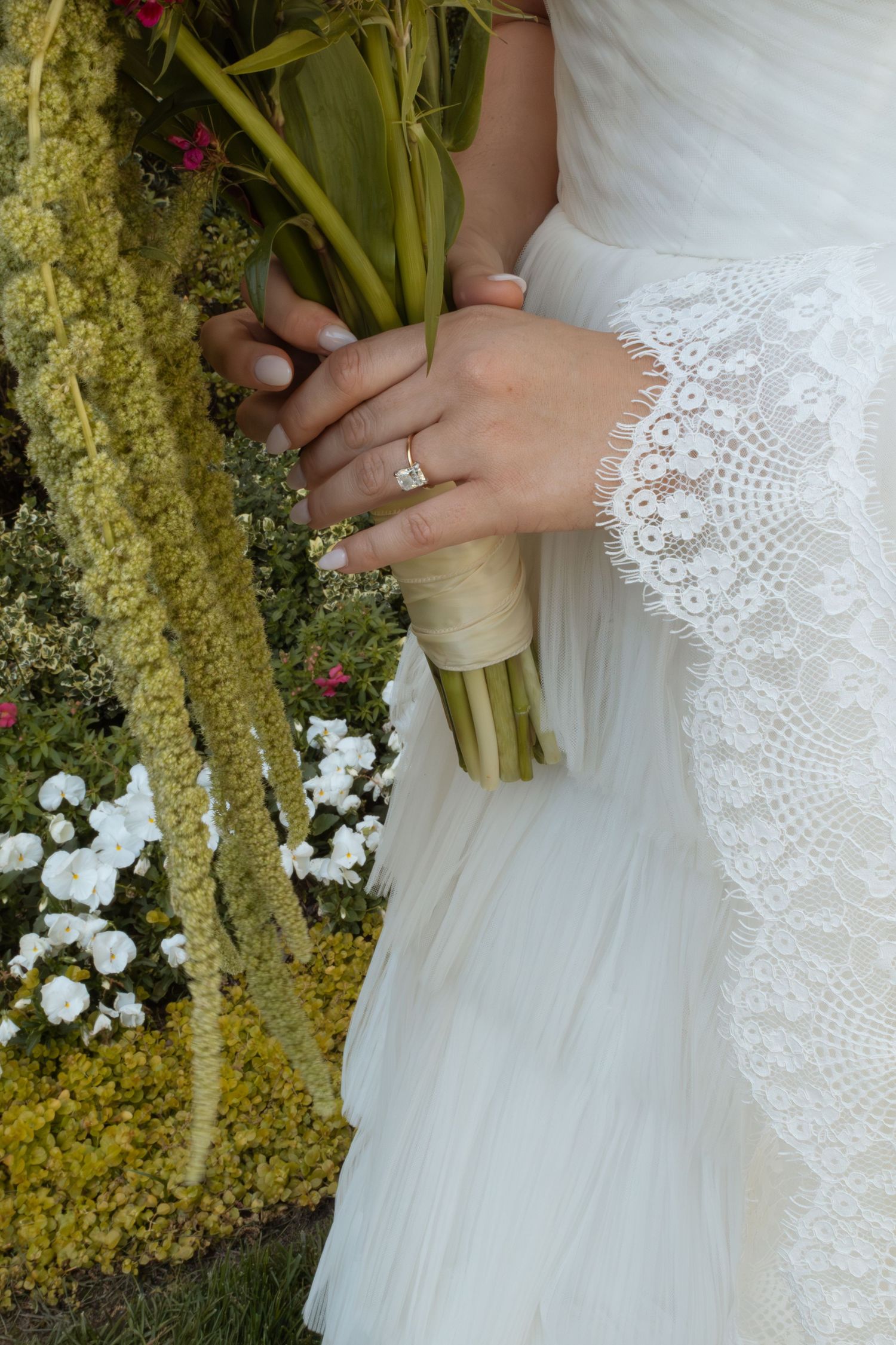 Bride in lace wedding dress holding a green bouquet with diamond engagement ring, standing near colorful garden flowers.