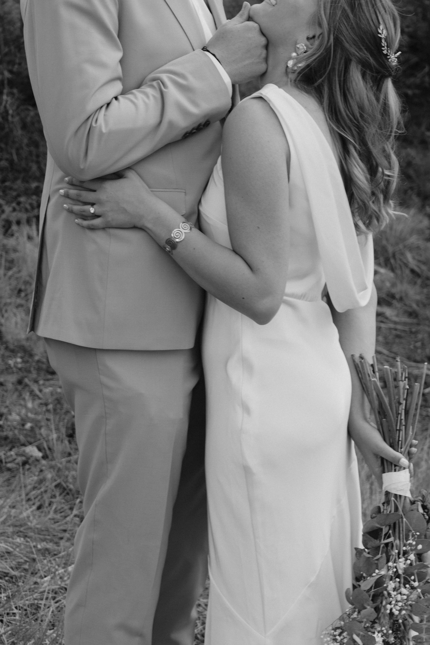 Black and white photo of bride and groom embracing outdoors, bride in white dress holding bouquet, groom in beige suit.