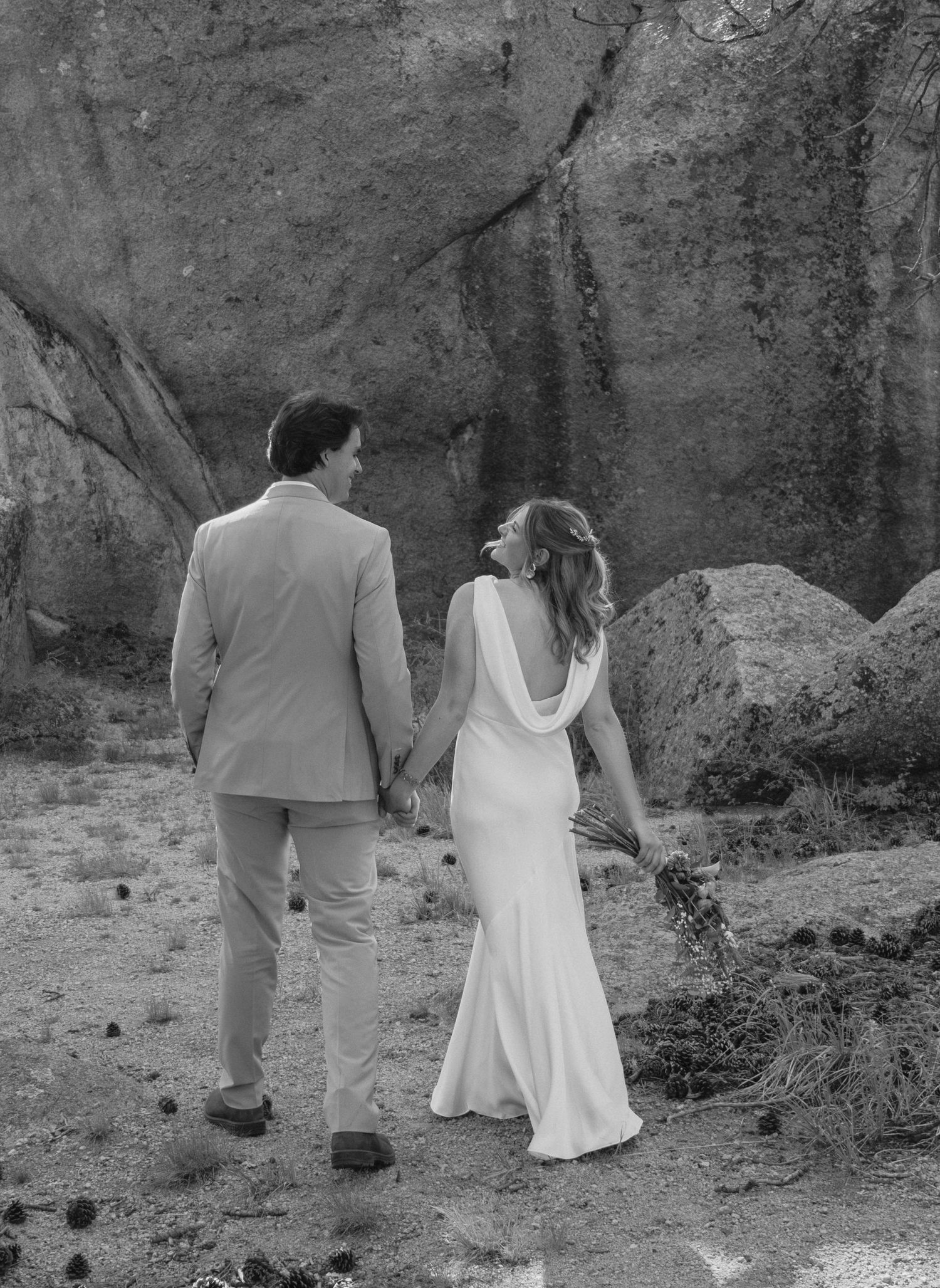 Black and white photo of a bride and groom holding hands walking toward large rocks on their wedding day.