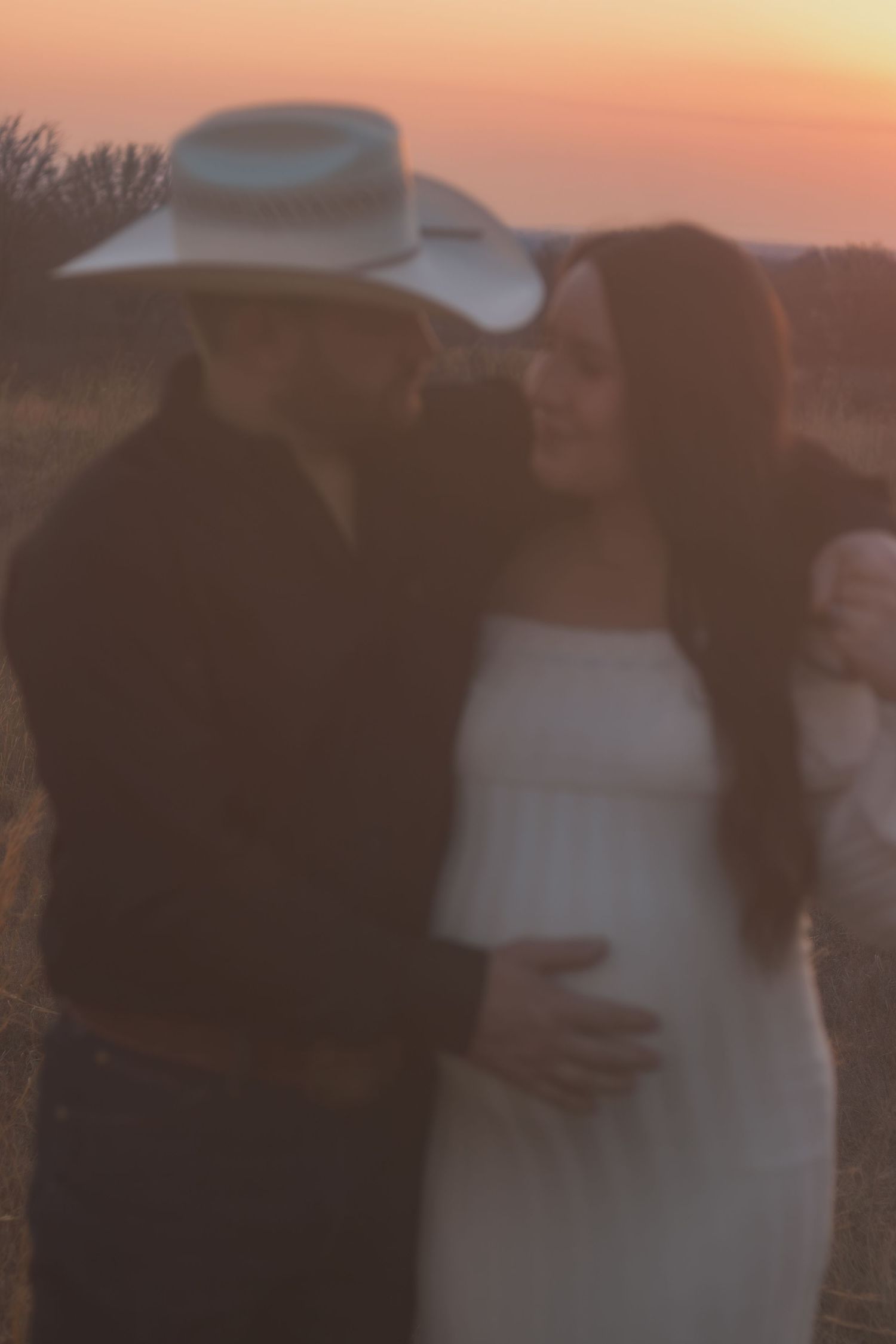 Couple embracing at sunset in a field, man wearing a white cowboy hat, woman in a white dress with a baby bump.