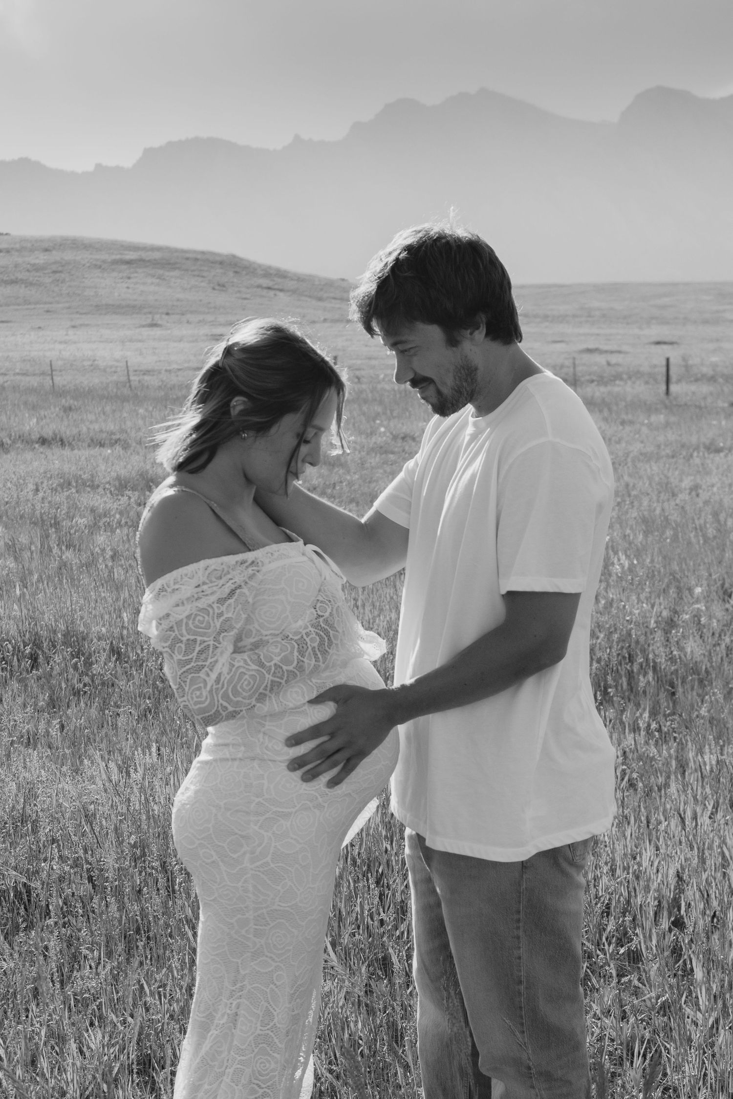 Black and white maternity photo of couple in field with mountains, man tenderly touching pregnant woman's belly.