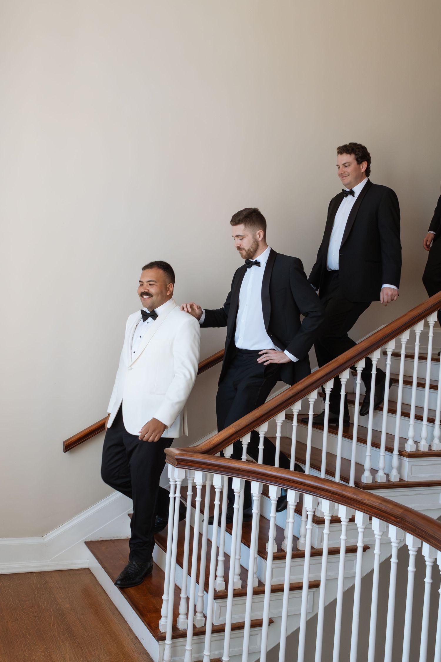 Three groomsmen in tuxedos descend an elegant curved staircase, with the groom in a white jacket leading.