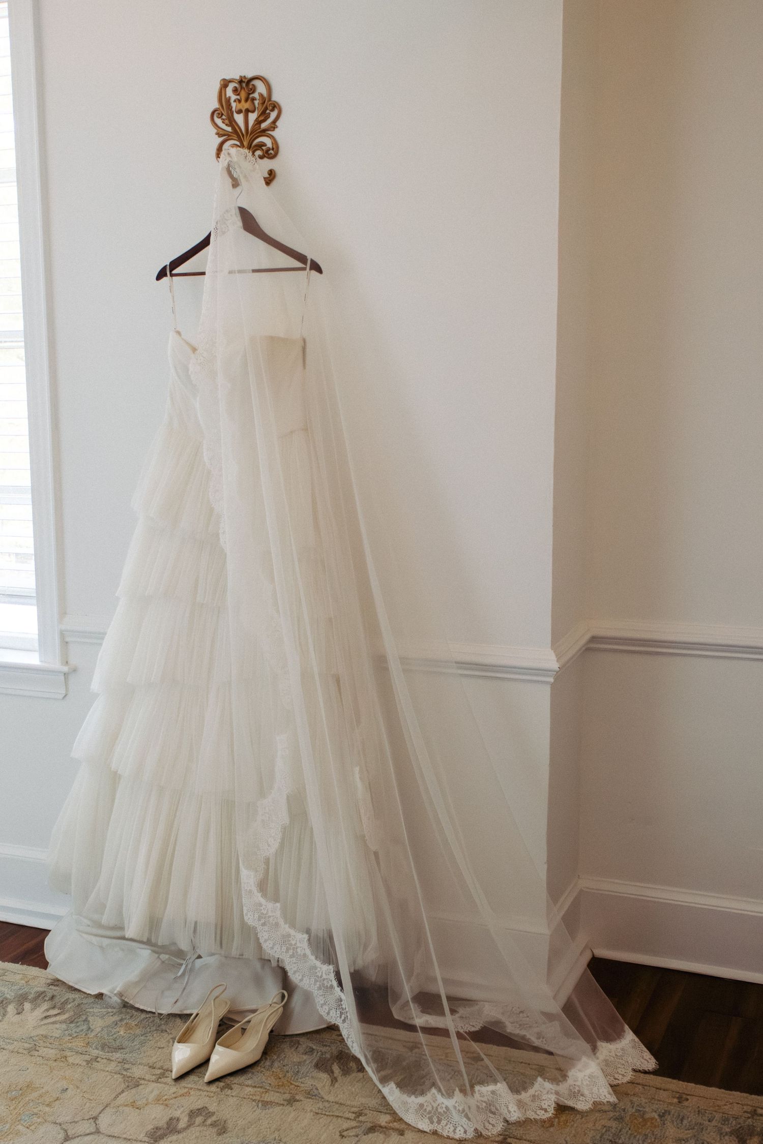 Elegant white tiered tulle wedding gown with lace-trimmed veil hanging on ornate hook beside bridal heels.