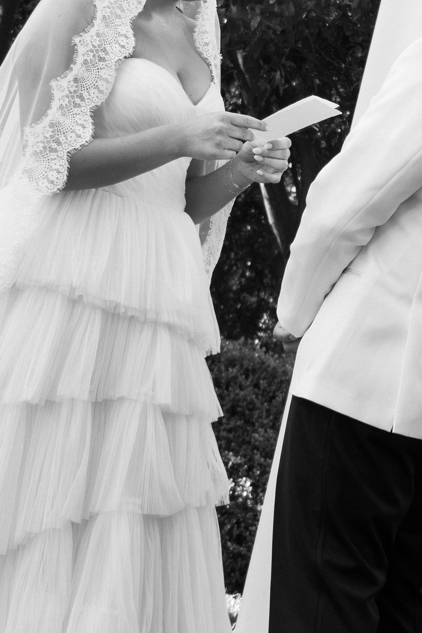 Black and white photo of a bride in tiered gown and veil reading wedding vows during outdoor ceremony.
