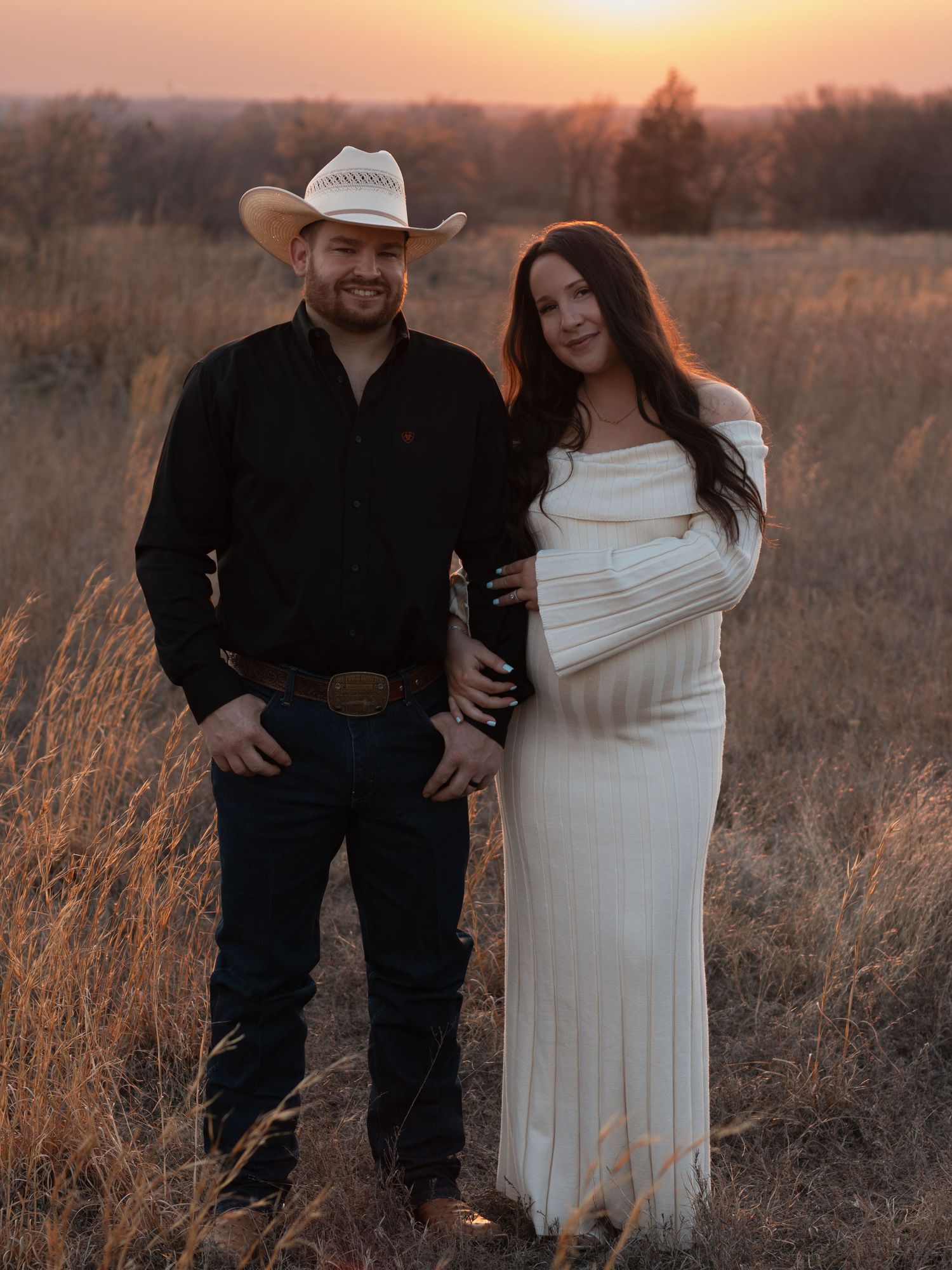 A couple poses together in a rustic field during golden hour, with her wearing white and him in black attire.
