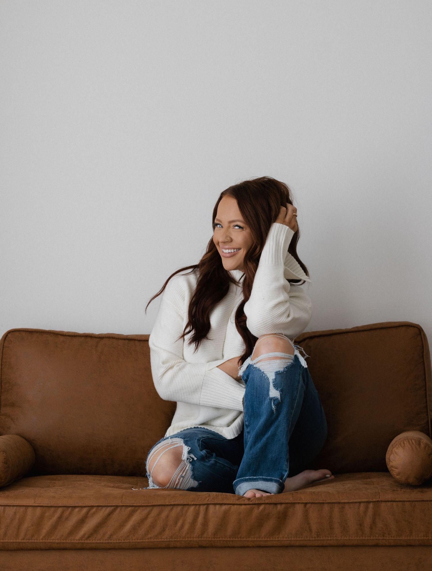 Smiling woman with dark hair in white sweater and ripped jeans sitting cross-legged on a brown leather couch.