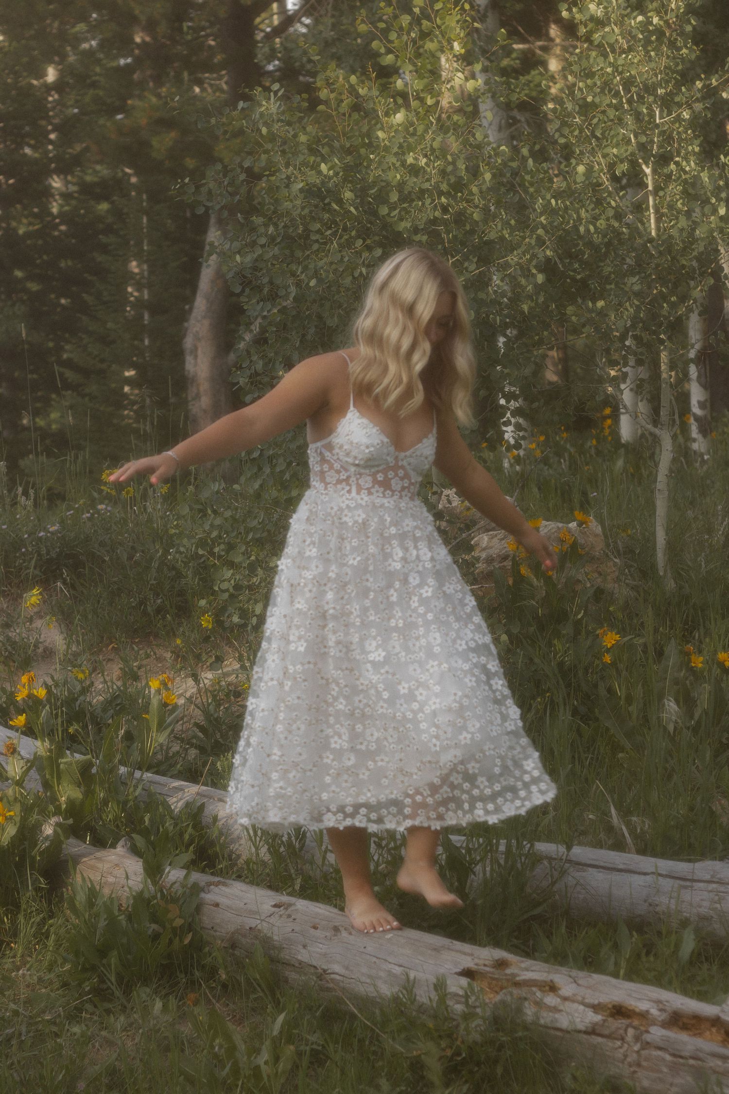 Blonde woman in white lace midi dress balancing barefoot on log surrounded by wildflowers in a forest.