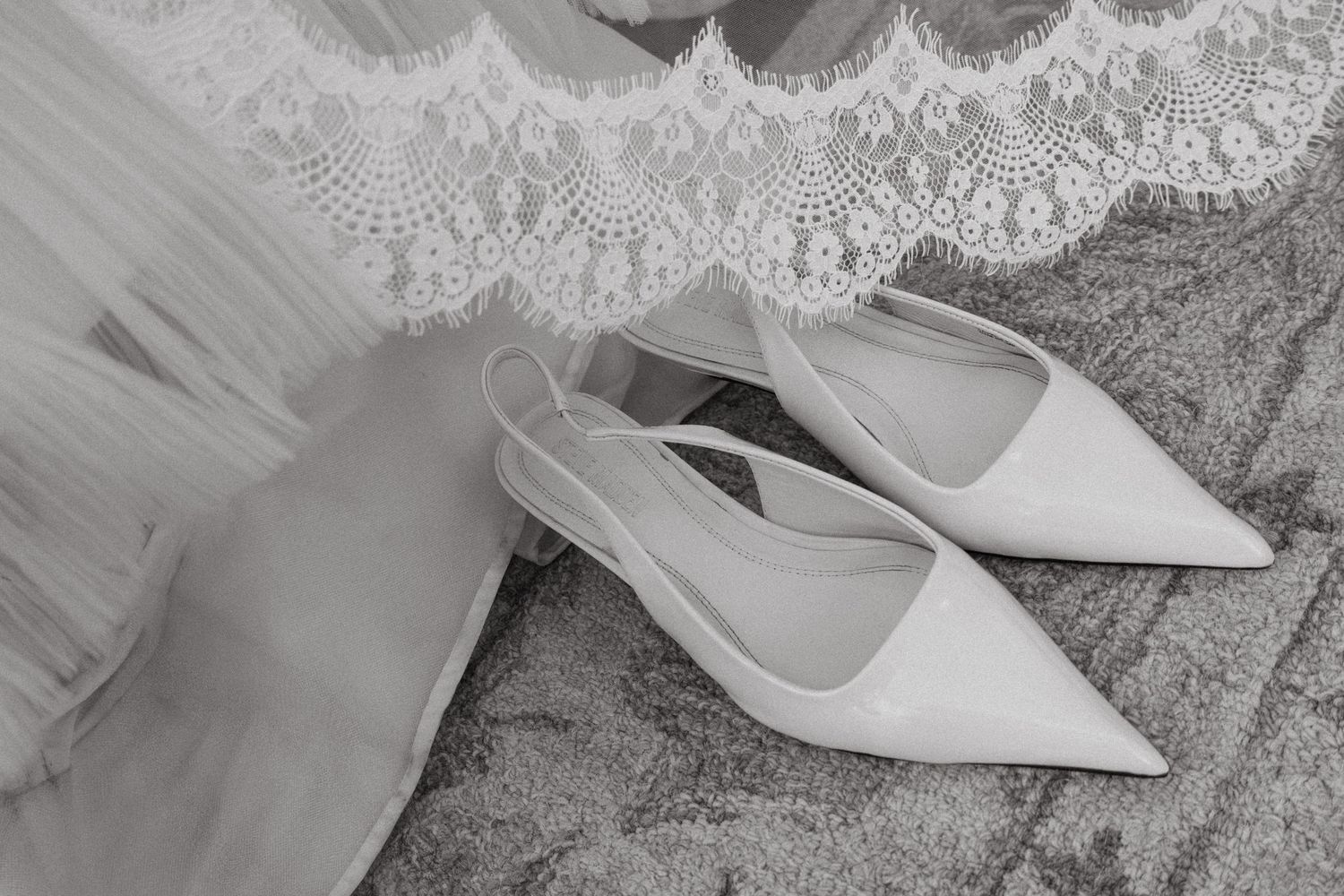 Elegant white bridal slingback heels with pointed toes beside a lace wedding dress, captured in black and white.
