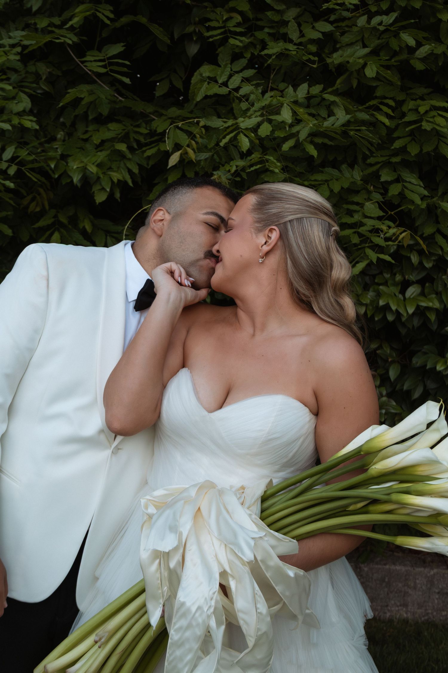 Bride and groom sharing a romantic kiss, bride holding white calla lily bouquet, groom in white tuxedo, lush green backdrop.