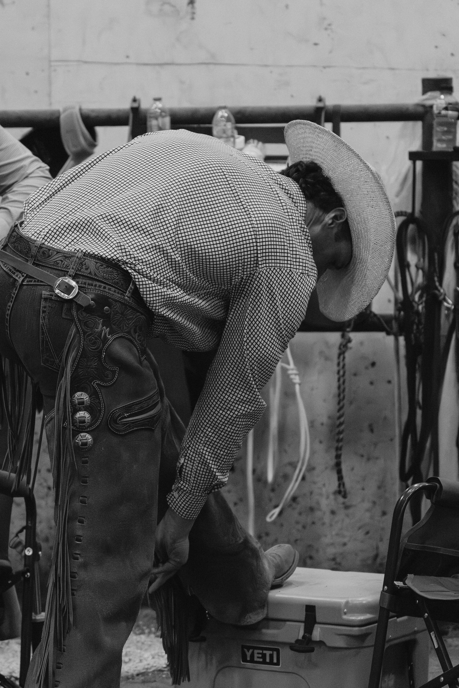 Black and white photo of a cowboy in chaps and hat bending over a Yeti cooler at a rodeo event.