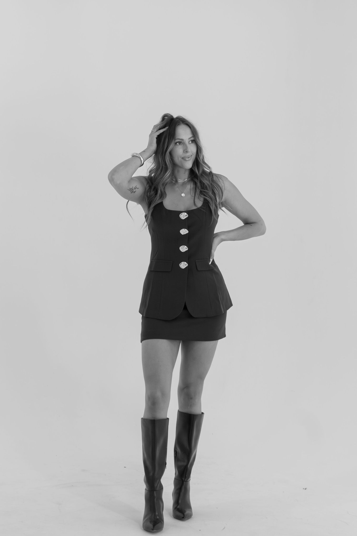 Person in black outfit and boots posing playfully against white background.