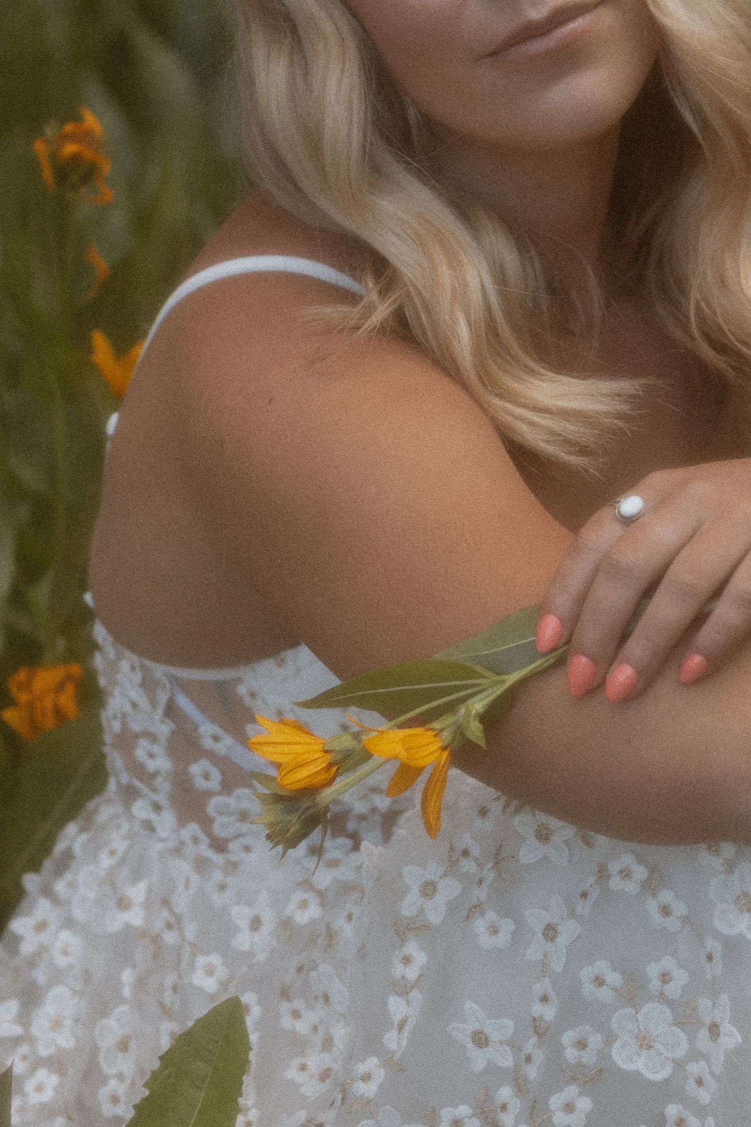 Blonde woman in white floral lace dress holding yellow wildflowers, with pink nails and a pearl ring.