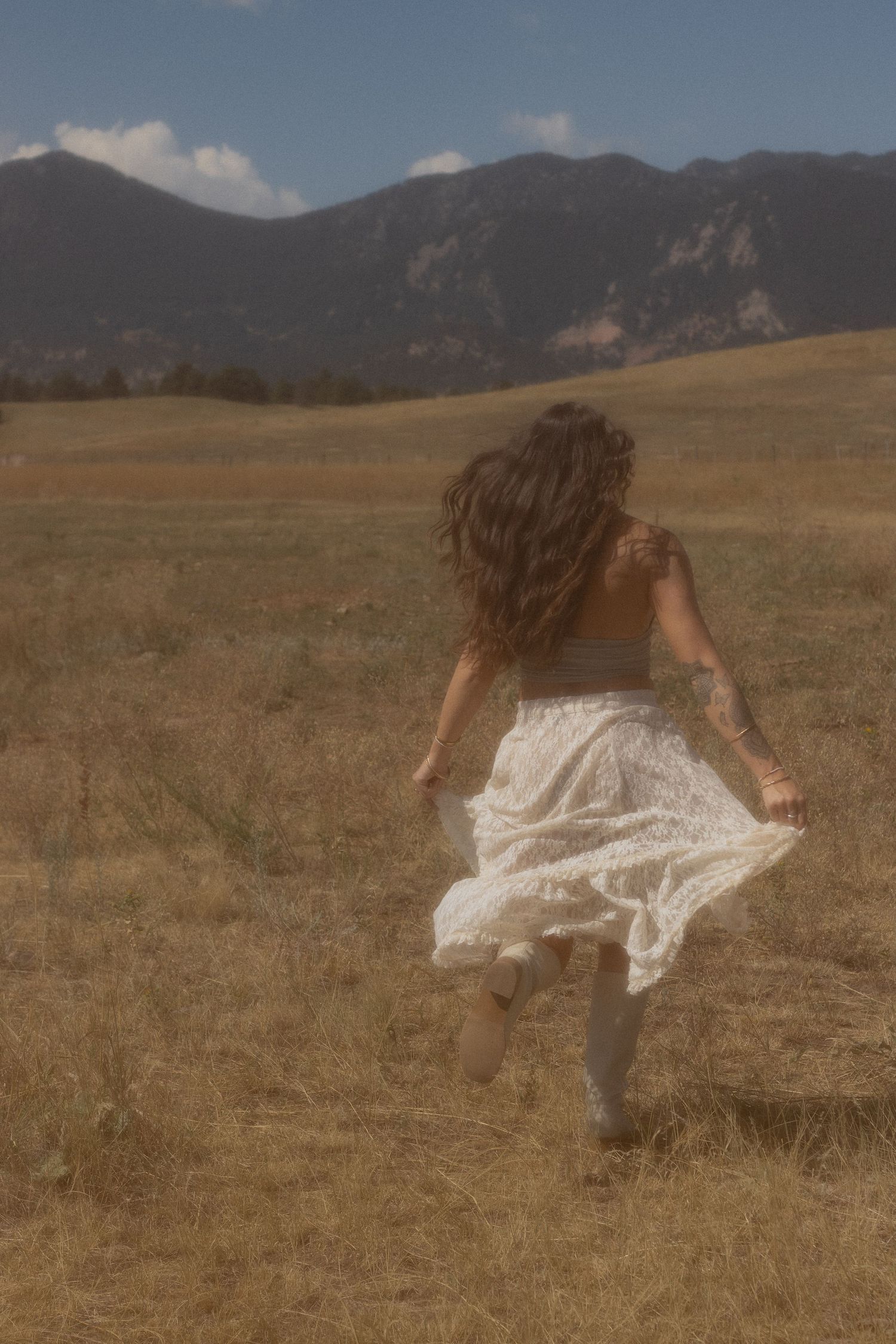 Person in white dress twirls gracefully across a sunlit meadow with mountains in the background.