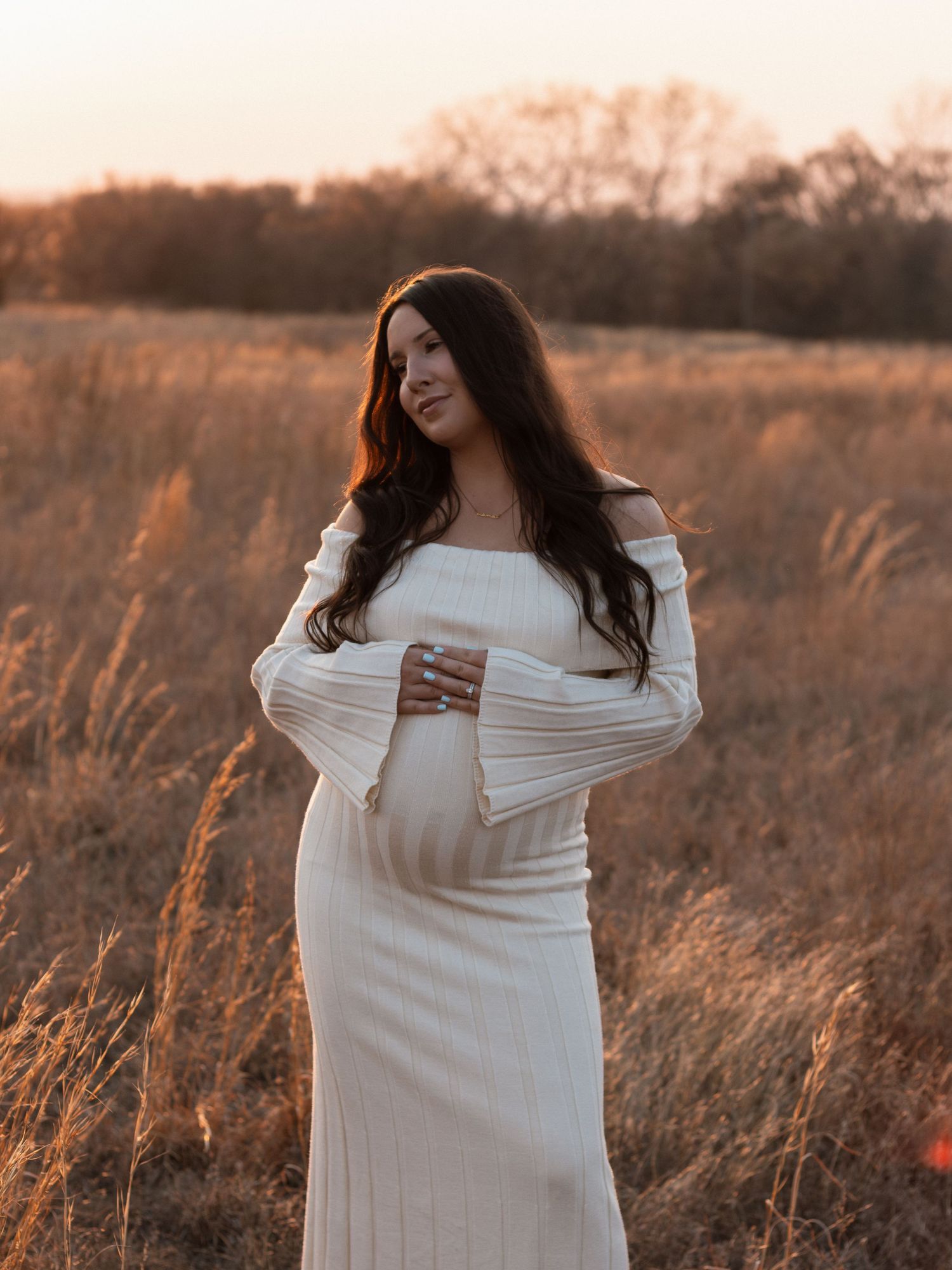 Pregnant woman in white off-shoulder dress standing in golden field at sunset, cradling baby bump.