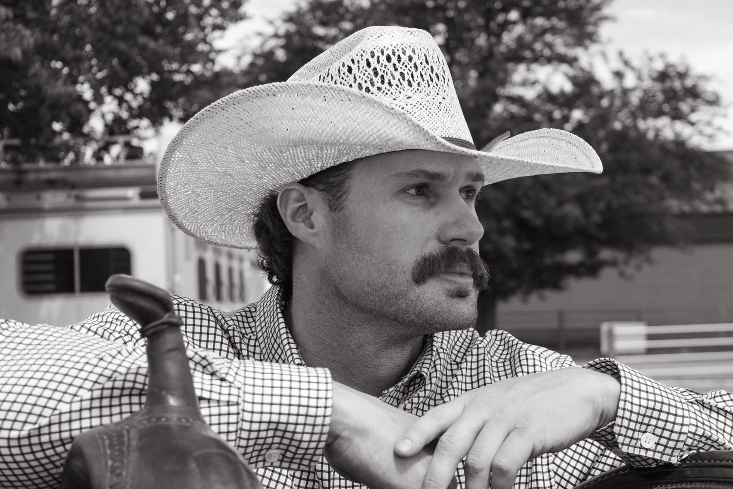 Black and white portrait of a man wearing a cowboy hat and checkered shirt, leaning on a fence outdoors.