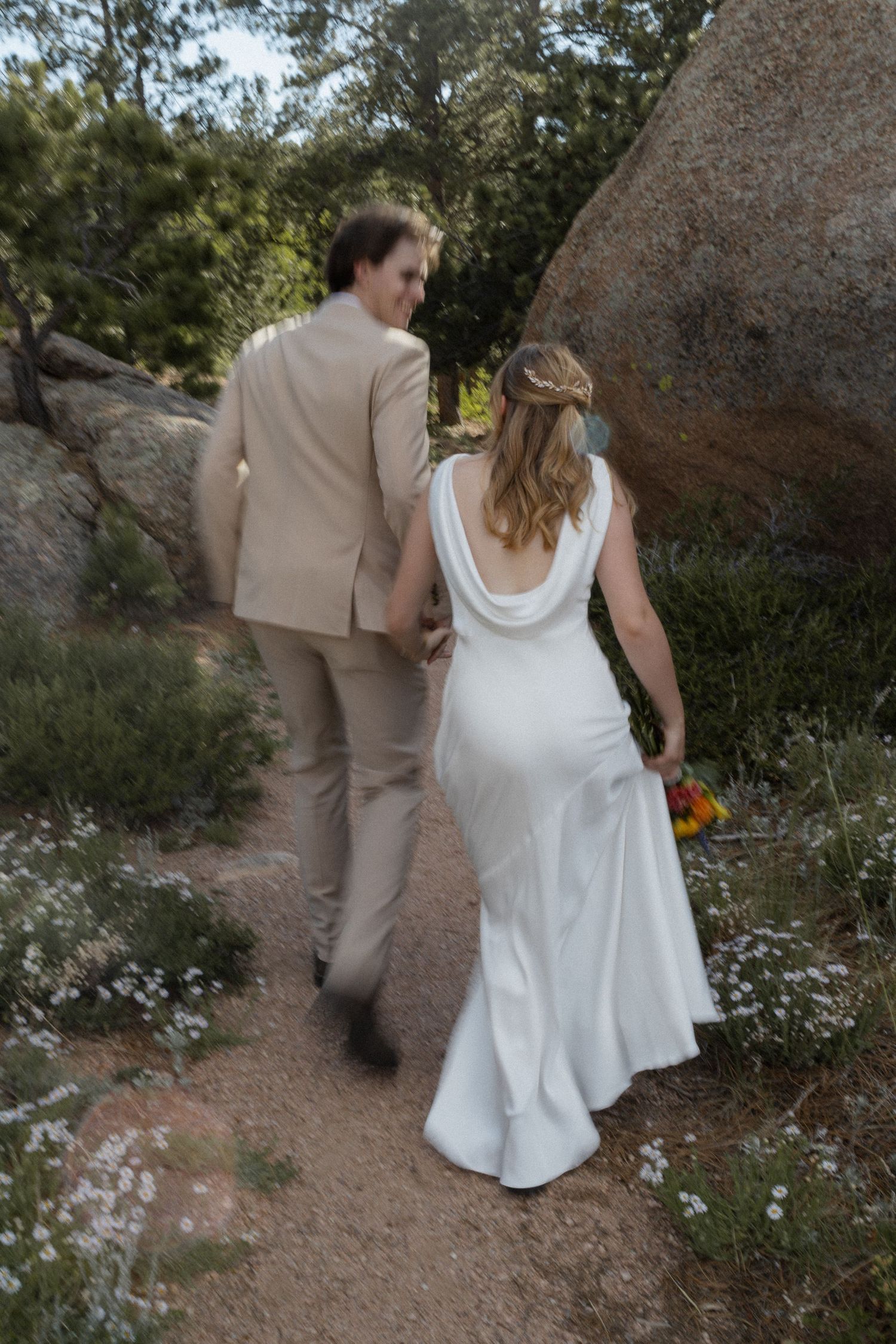 Bride and groom walking hand in hand on a garden path, bride in white backless gown holding colorful bouquet.