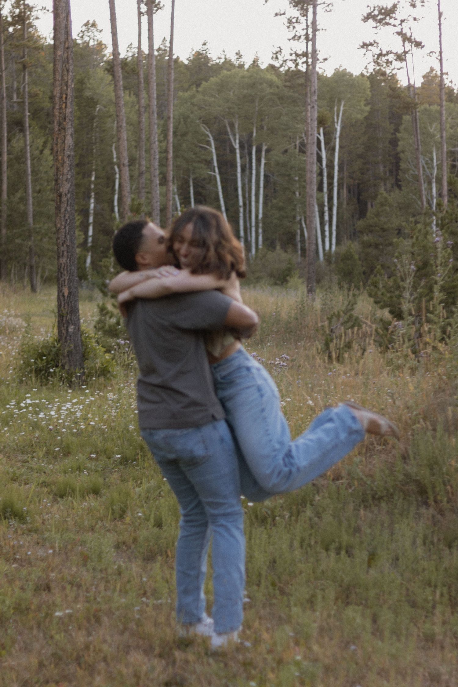 Playful couple spinning and embracing among pine trees in a forest setting.
