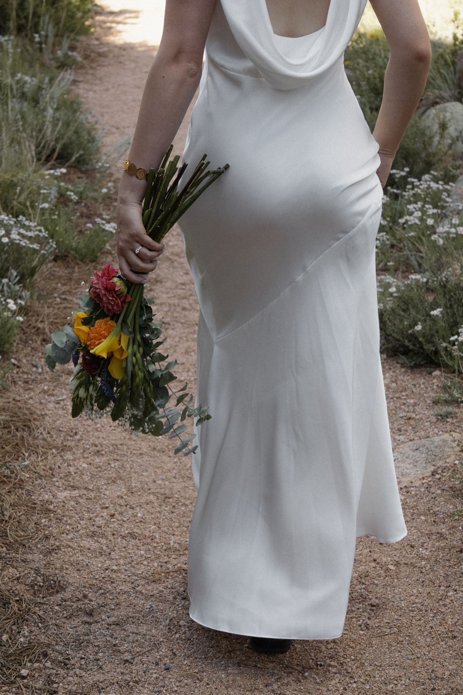 Bride in white slip dress holding colorful bouquet of flowers while walking on sandy path outdoors.