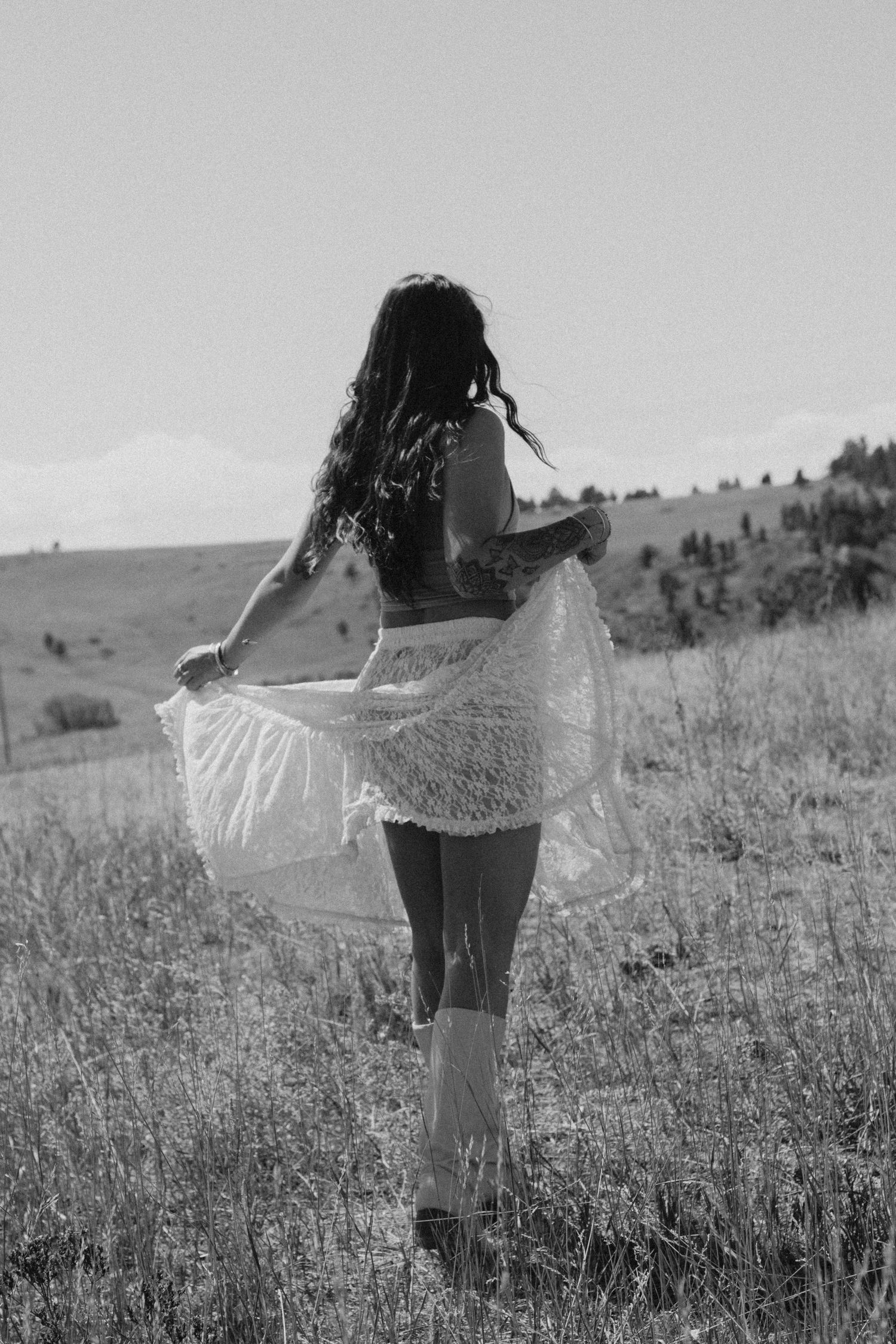 Black and white photo of a woman in lace skirt and boots standing in a field, viewed from behind.