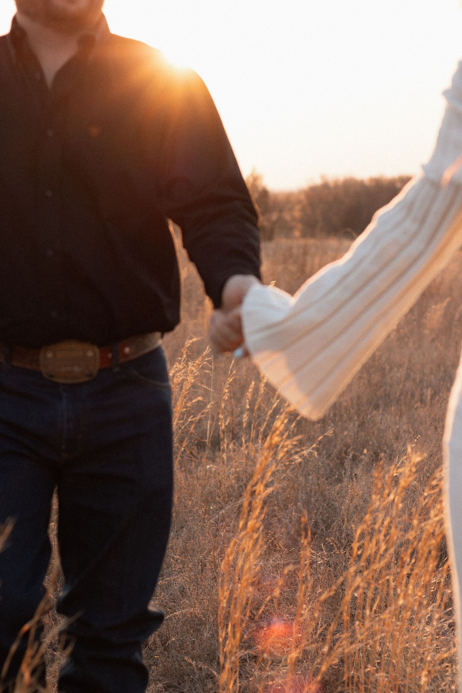 Couple holding hands at golden sunset in a field, man in dark shirt and jeans, woman in white sweater.