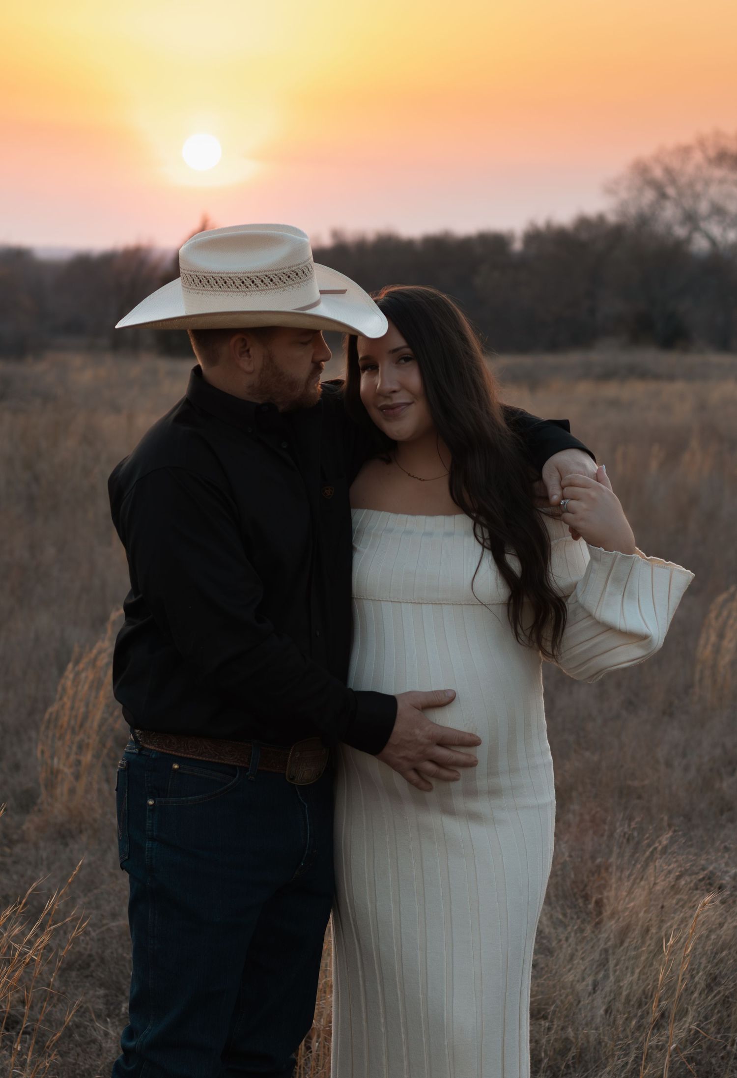 Couple poses in golden field at sunset, man in cowboy hat embraces pregnant woman in white dress.