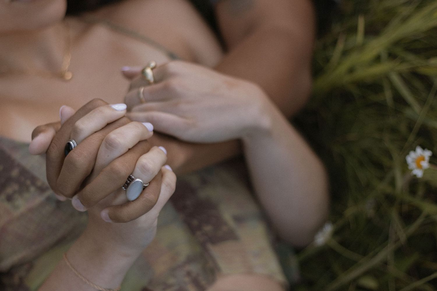Two people holding hands in grass, wearing silver and gold rings, with white manicured nails and a daisy nearby.