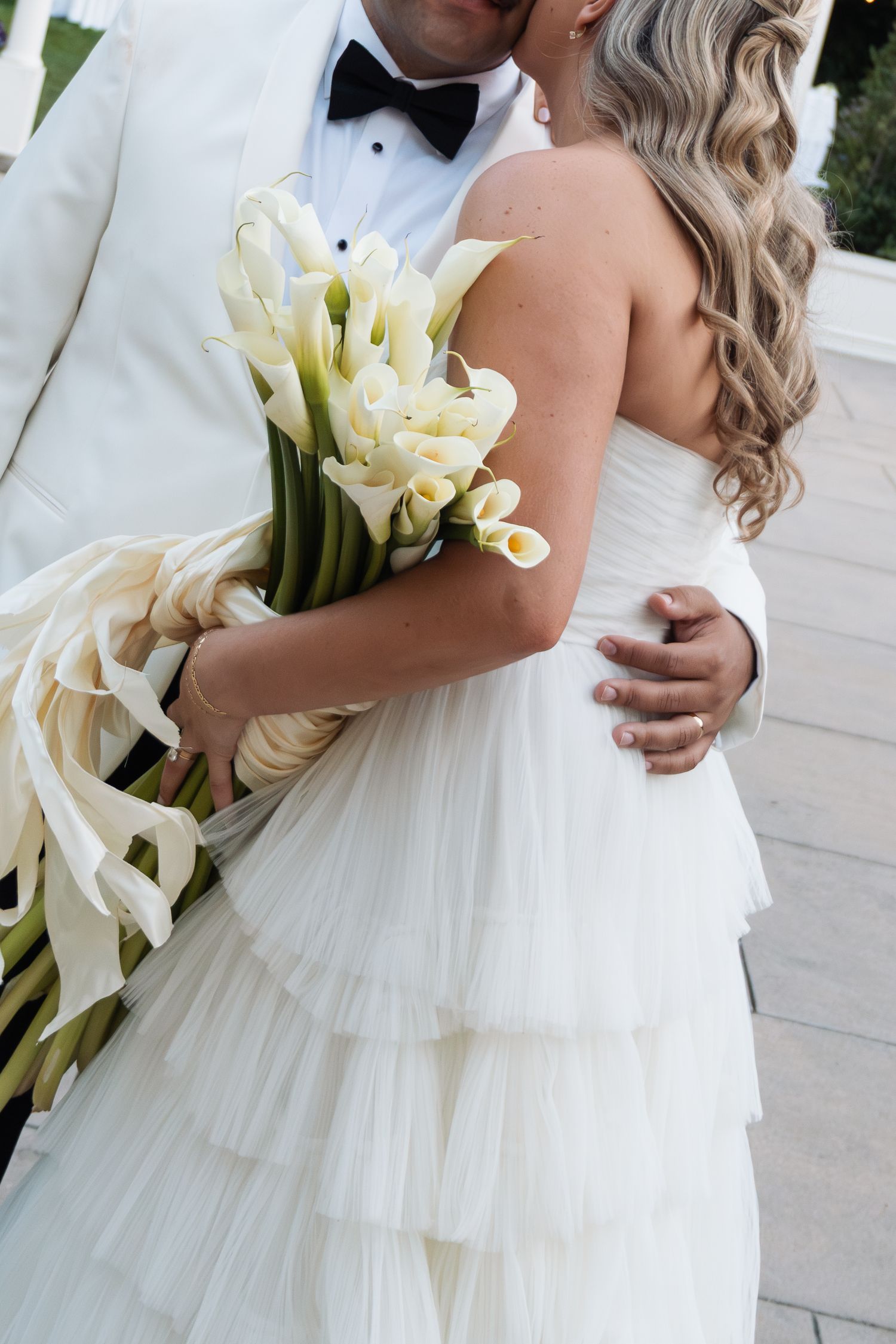 Bride and groom embracing, bride holding white calla lily bouquet, wearing strapless tiered gown, groom in white tuxedo.