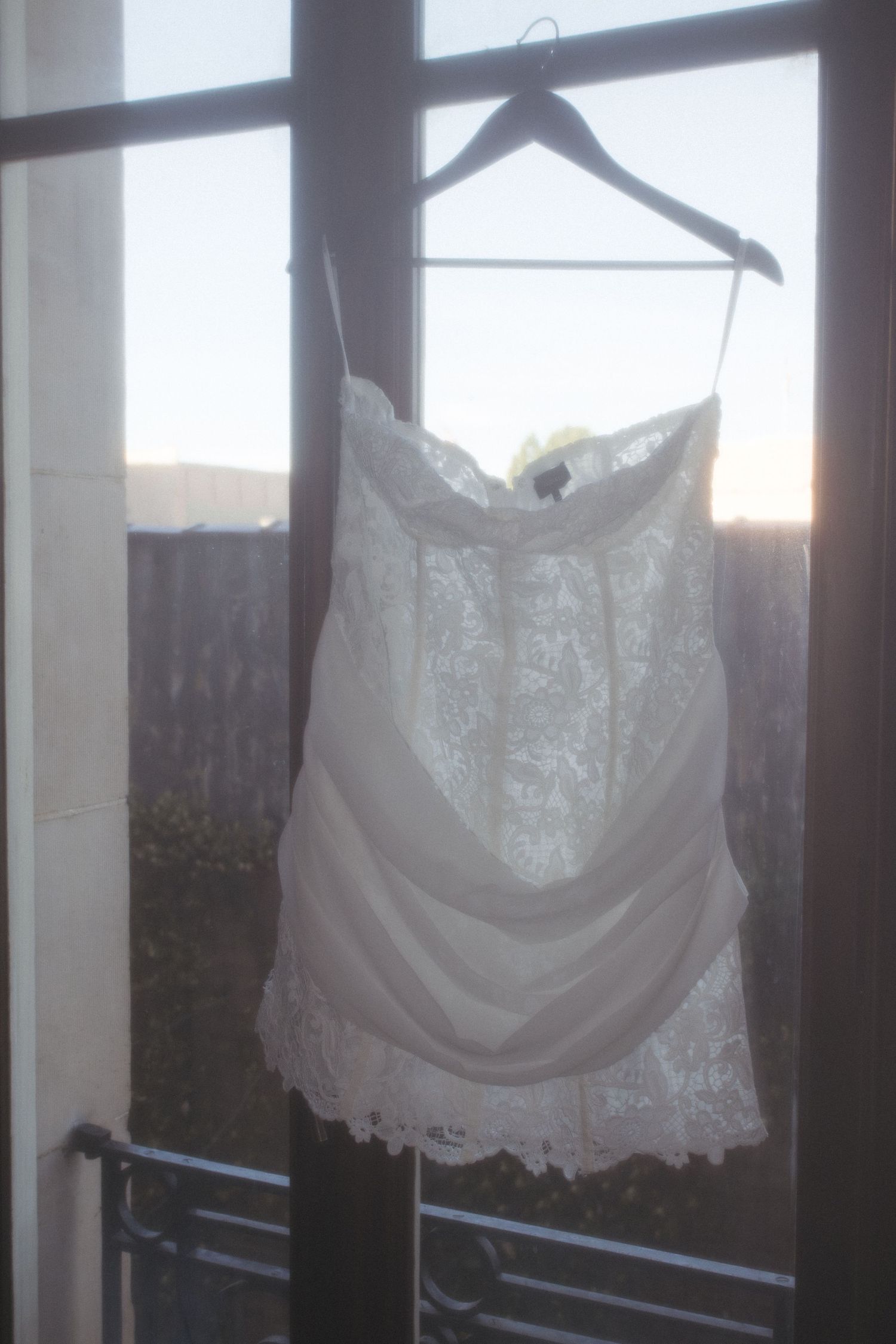 White lace camisole top hanging on a wooden hanger in front of a window at sunset.