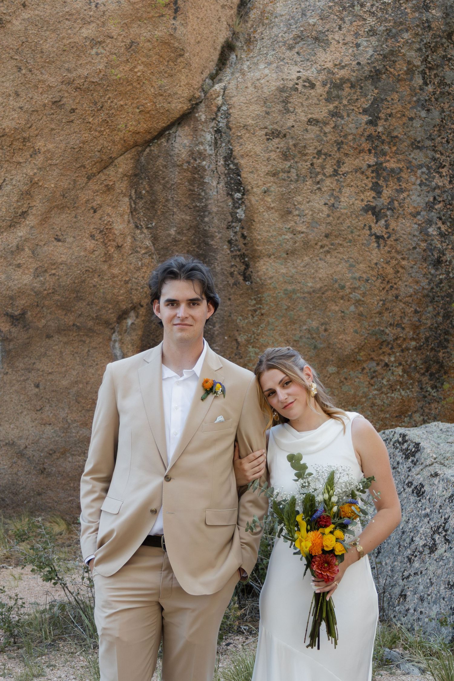Bride and groom posing before large rock, bride in white dress with colorful bouquet, groom in beige suit.