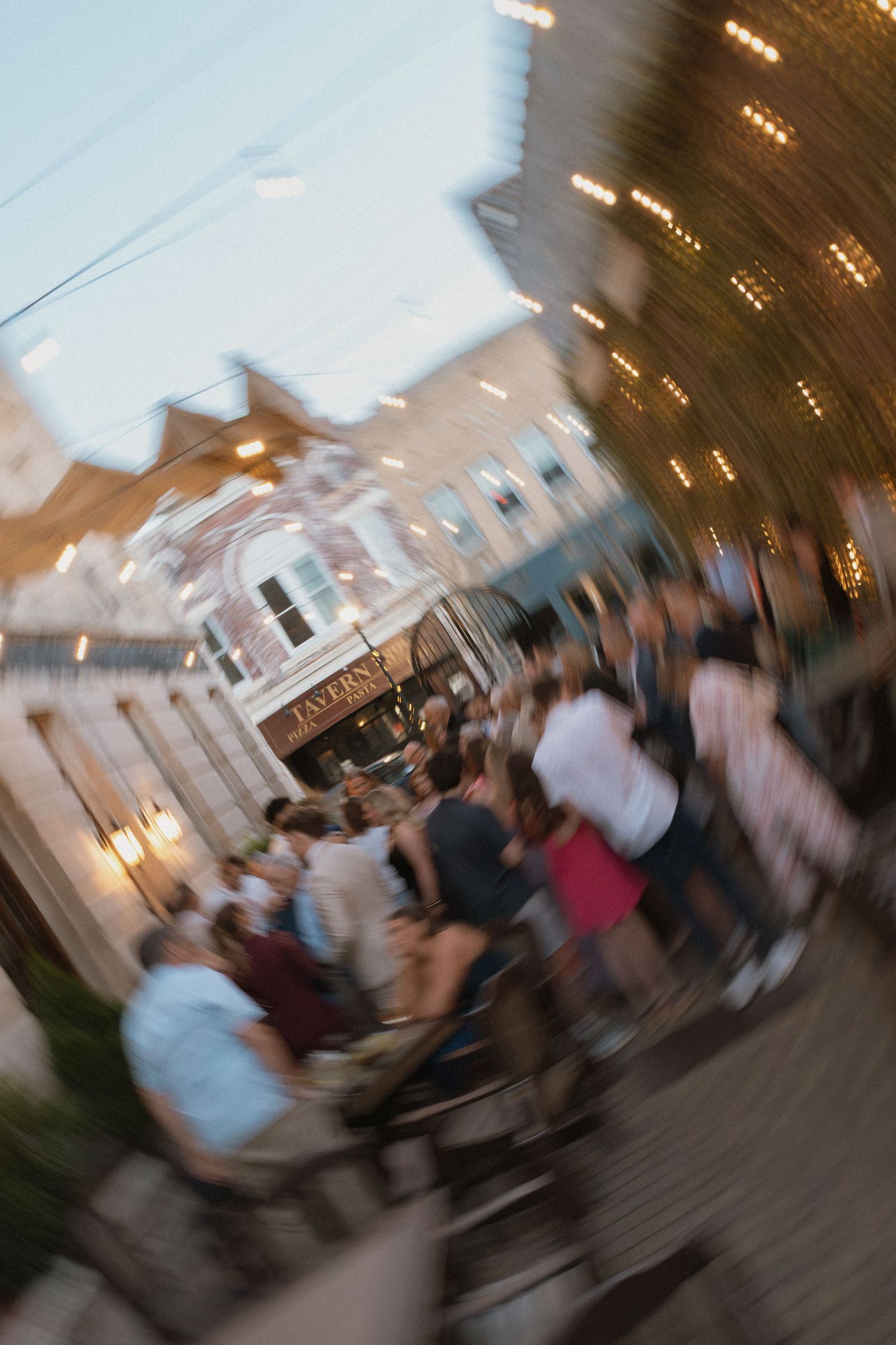 Blurred motion photo of a lively outdoor crowd gathered near a tavern at dusk with string lights.