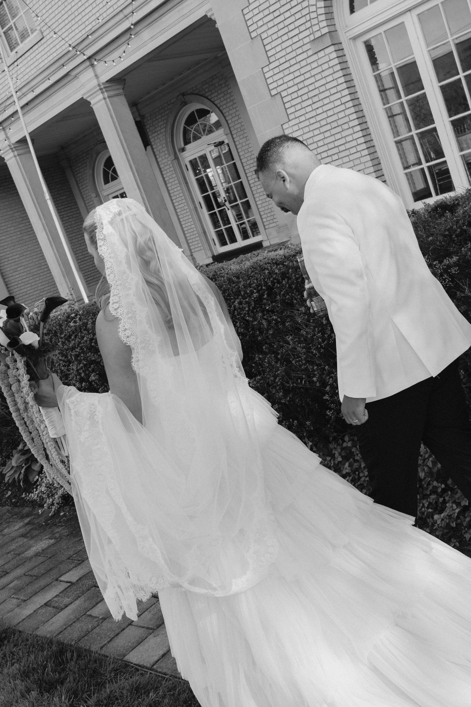 Black and white photo of a bride in a flowing gown and veil walking with a groom in a white tuxedo jacket.