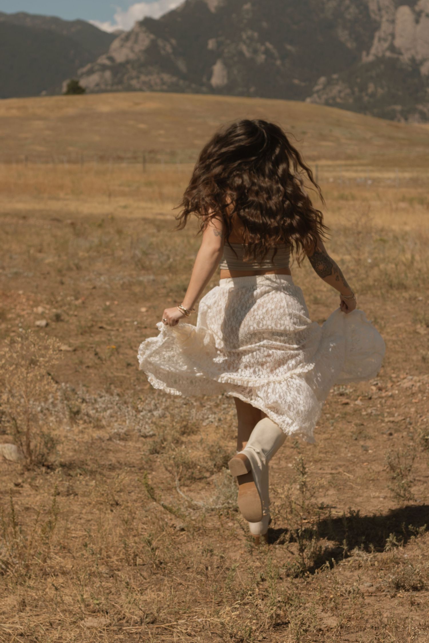 A woman in a white lace skirt runs through a golden field with mountains in the background.
