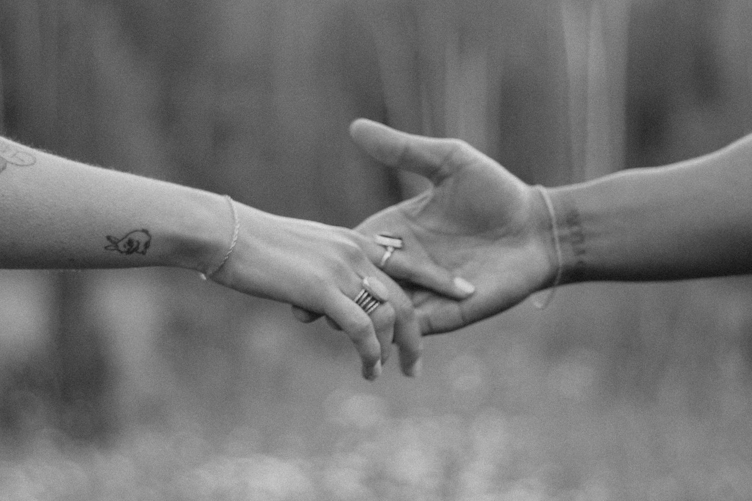 Black and white photo of two people gently holding hands, one with a small tattoo and delicate bracelet.