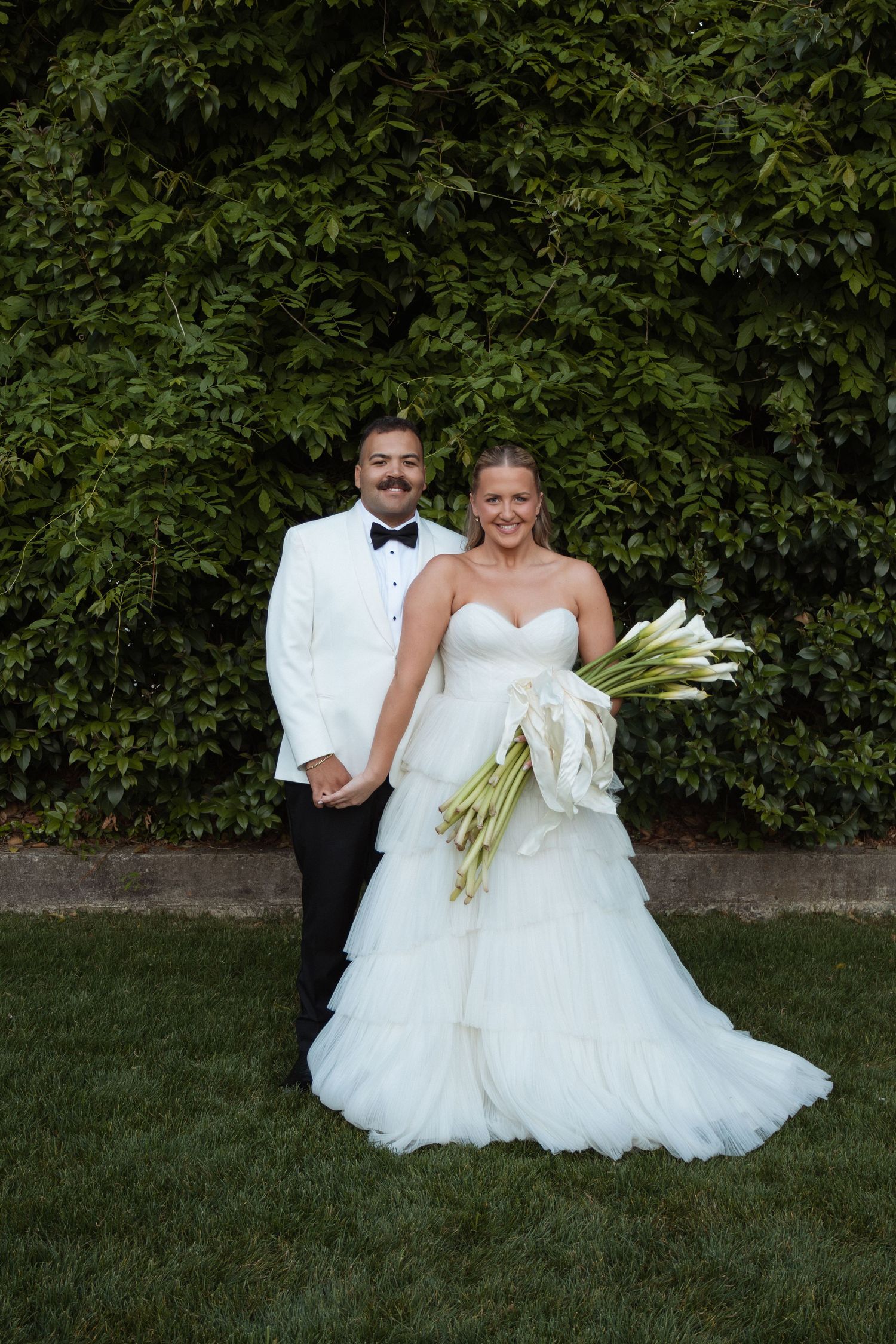 Bride in white strapless gown holding calla lily bouquet stands with groom in white tuxedo jacket on green lawn.