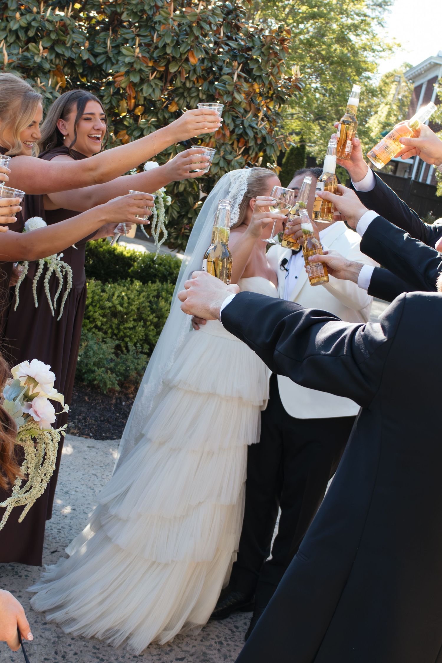 Wedding party raising glasses in a champagne toast, bride in white gown and veil surrounded by bridesmaids and groomsmen outdoors.