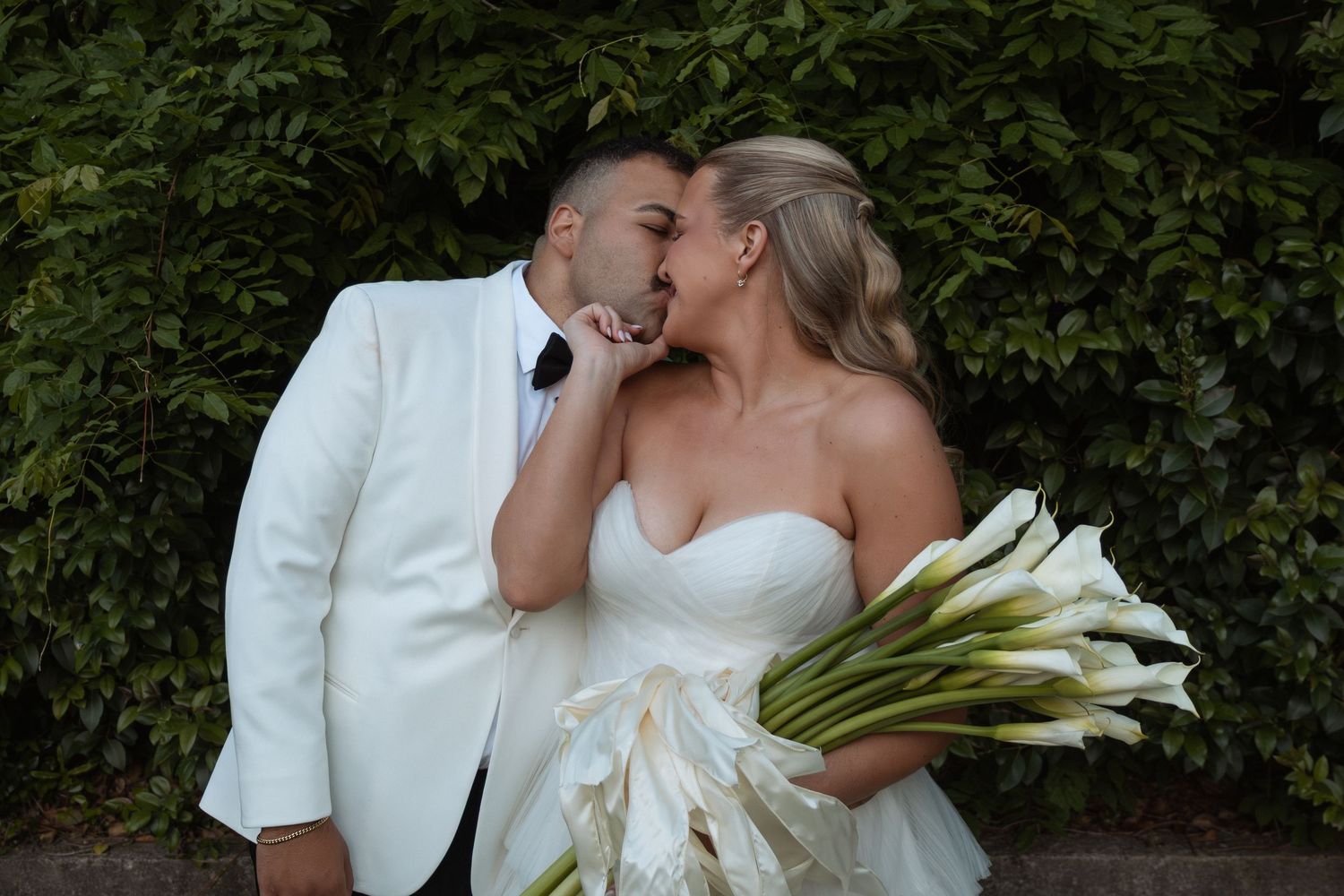 Bride and groom share a romantic kiss, she holds white calla lilies bouquet, he wears white tuxedo, green foliage backdrop.