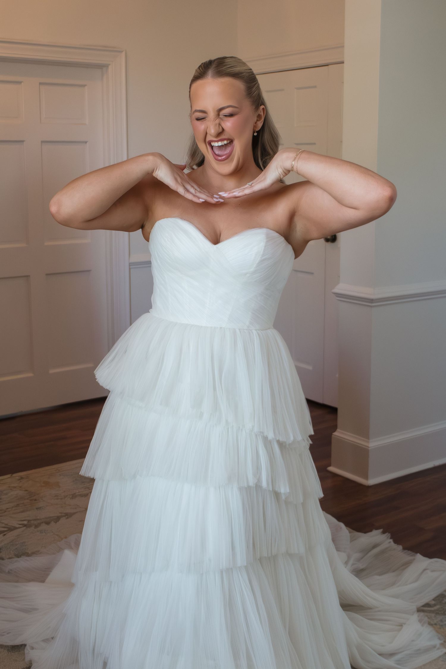 Smiling bride in a strapless white tiered tulle ball gown wedding dress, adjusting her necklace indoors.