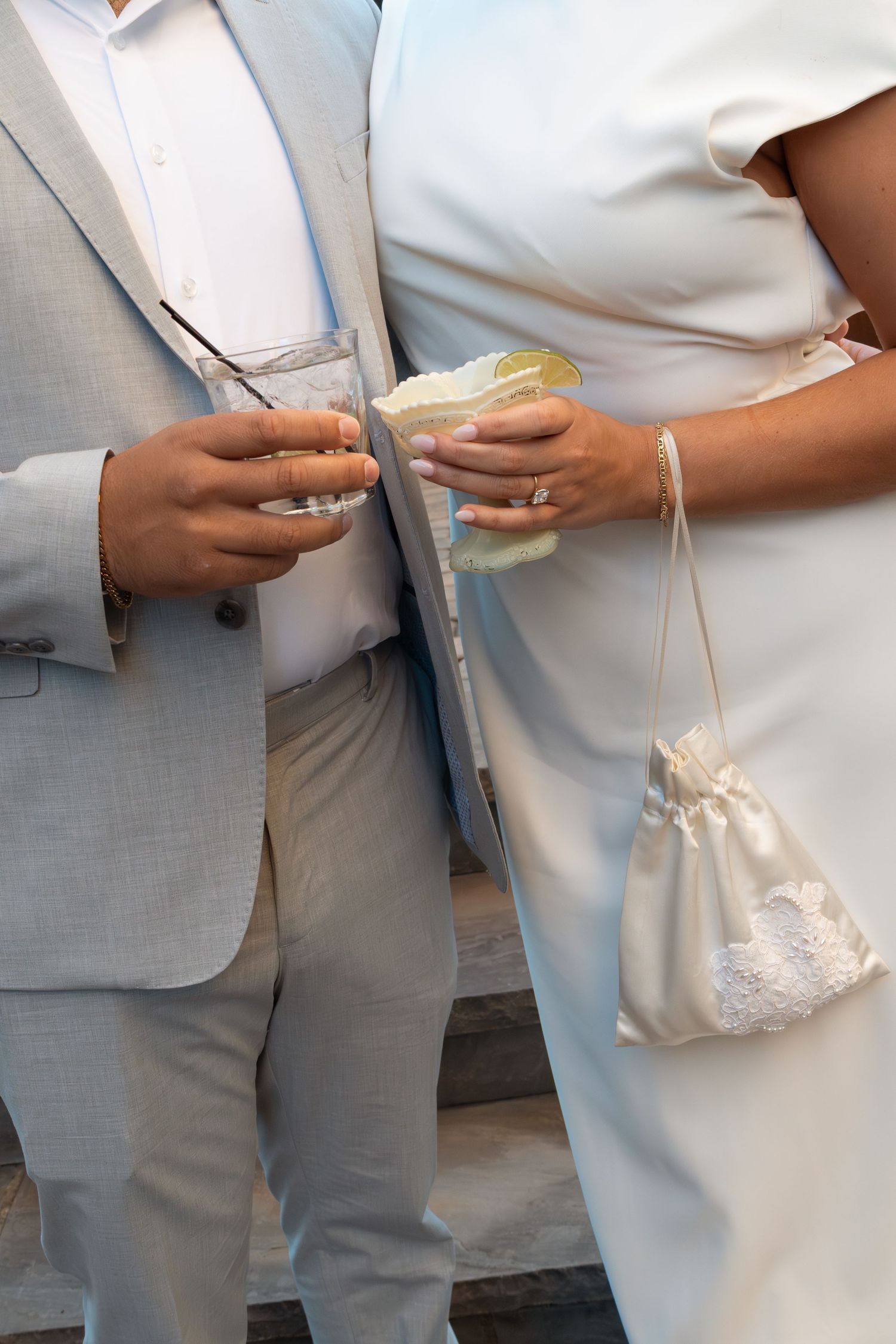 Bride and groom holding drinks at wedding, woman wearing white dress with satin purse and diamond ring.