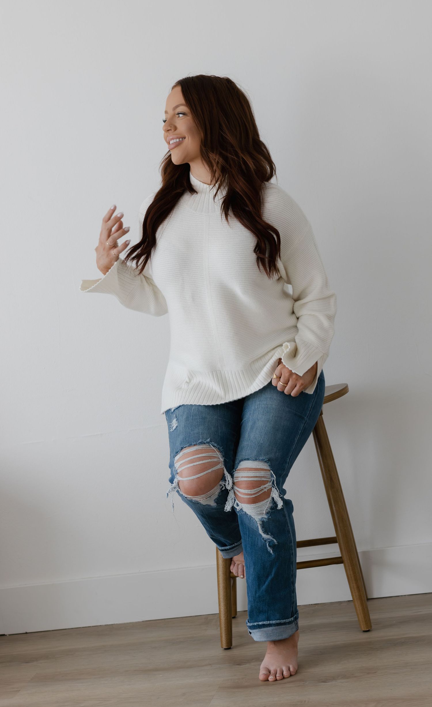 Woman in cream oversized sweater and distressed jeans sits on wooden stool, smiling and barefoot against white wall.