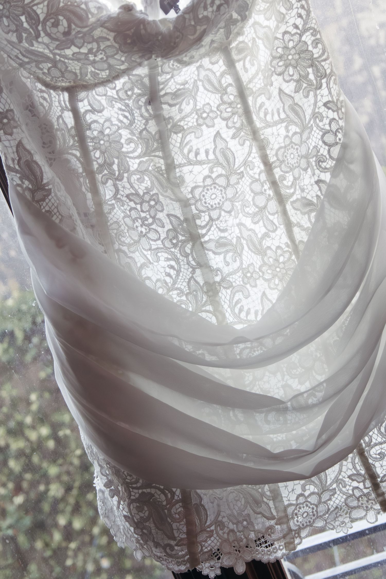 Elegant white lace curtain with floral pattern draped over a sheer fabric panel in a sunlit window.