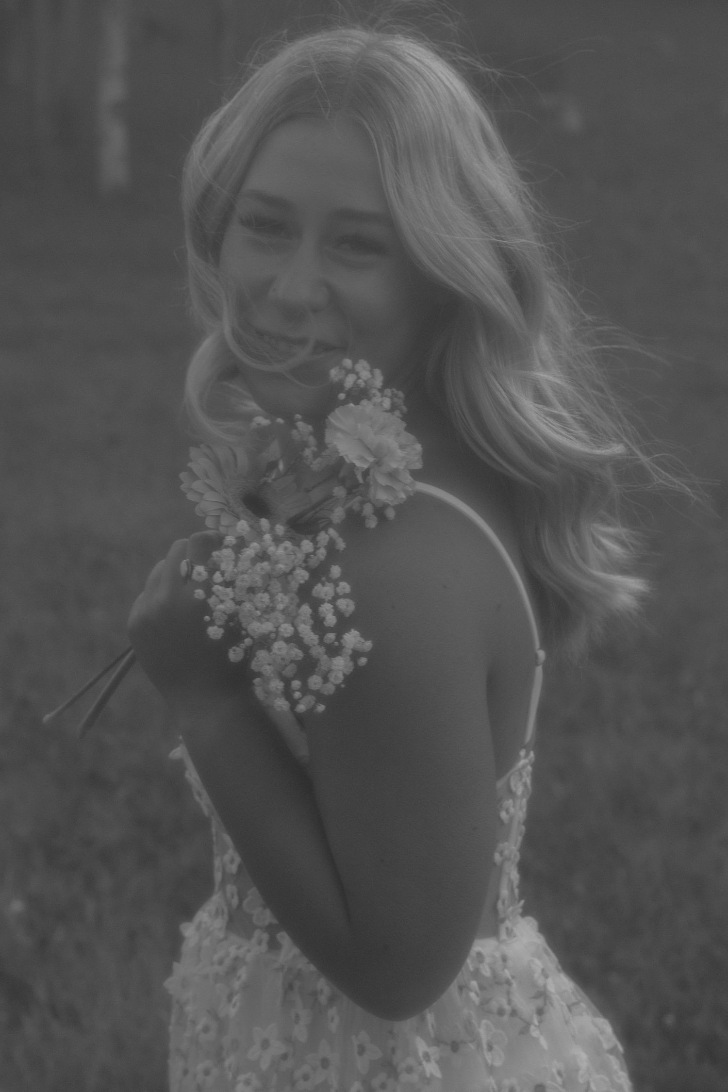 Smiling blonde woman in floral dress holding bouquet of wildflowers, black and white portrait photography.
