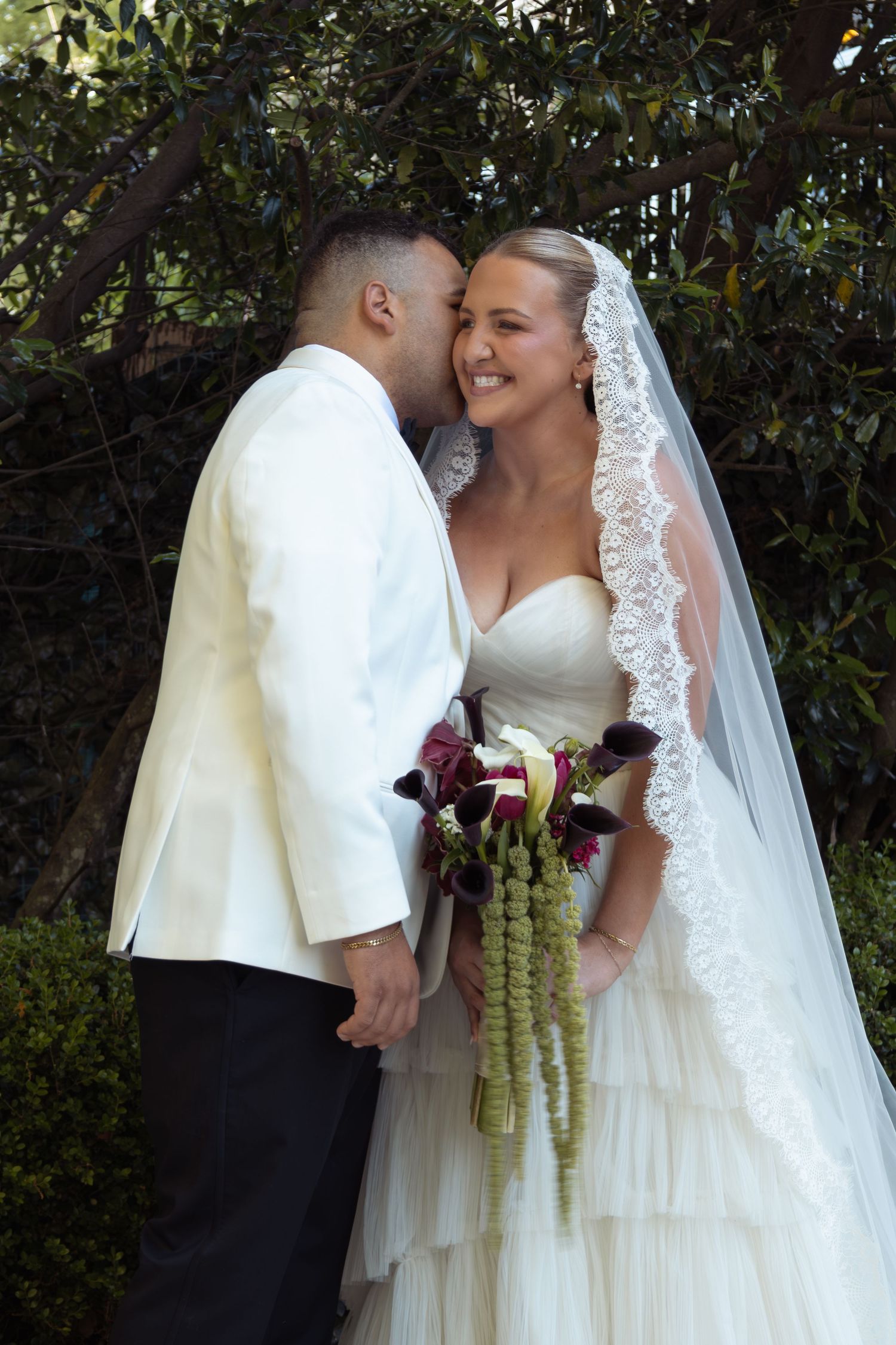 Bride in white gown and veil holds bouquet as groom in white jacket whispers in her ear outdoors.