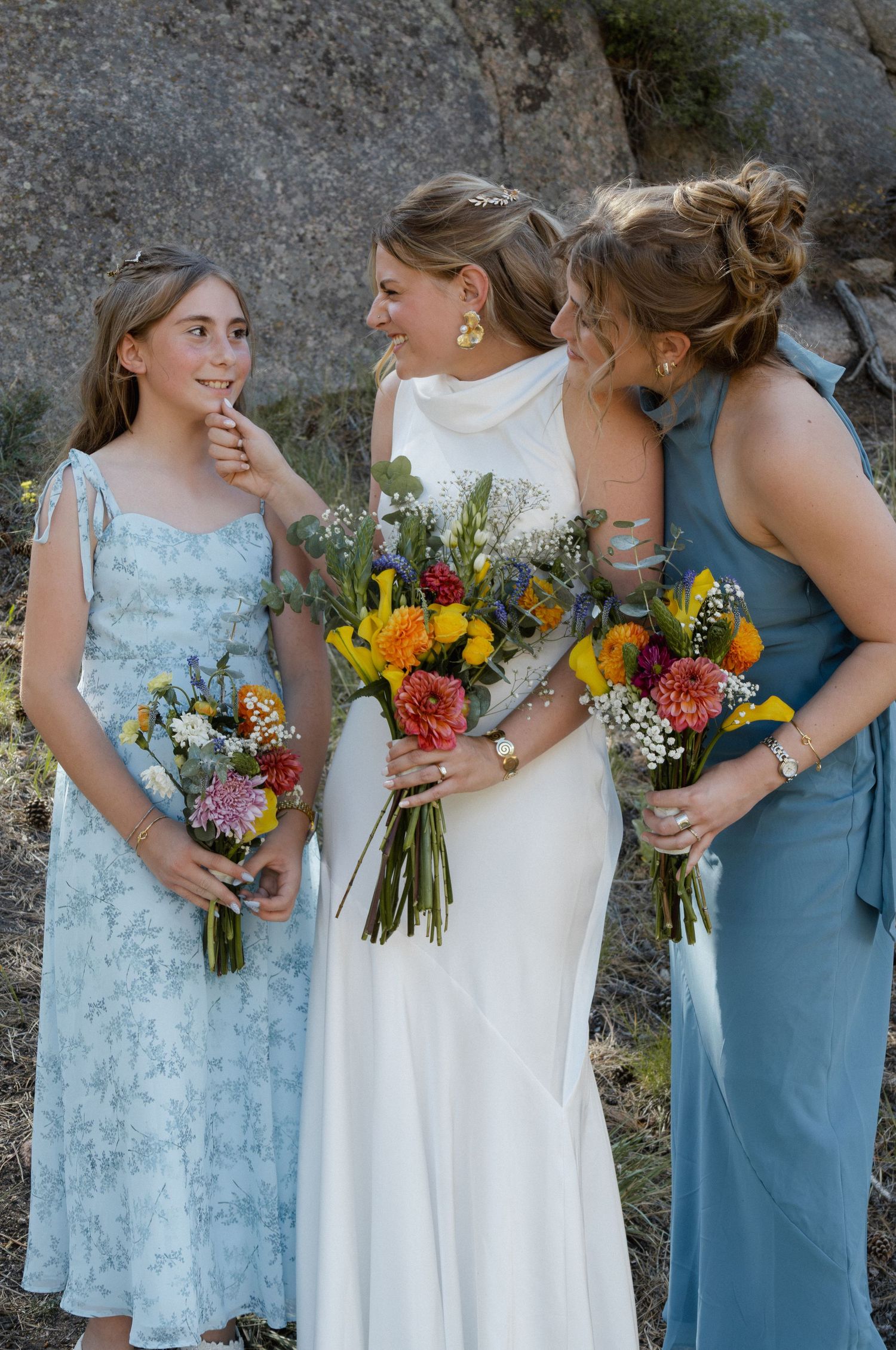 Bride in white gown with bridesmaids holding colorful wildflower bouquets, smiling together outdoors near rocky backdrop.