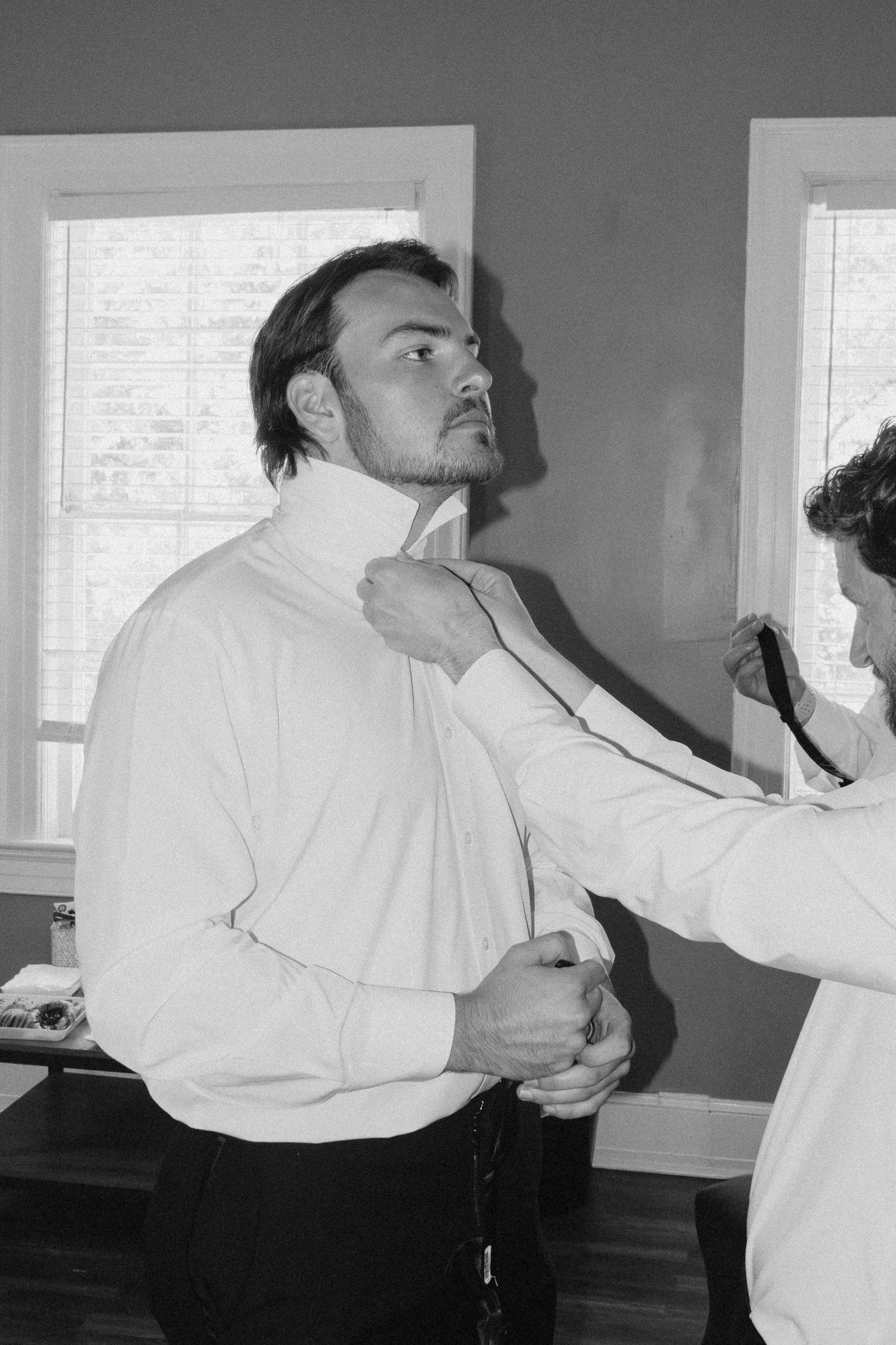 Black and white photo of a man in a white dress shirt being helped with his tie while getting ready for a formal event.