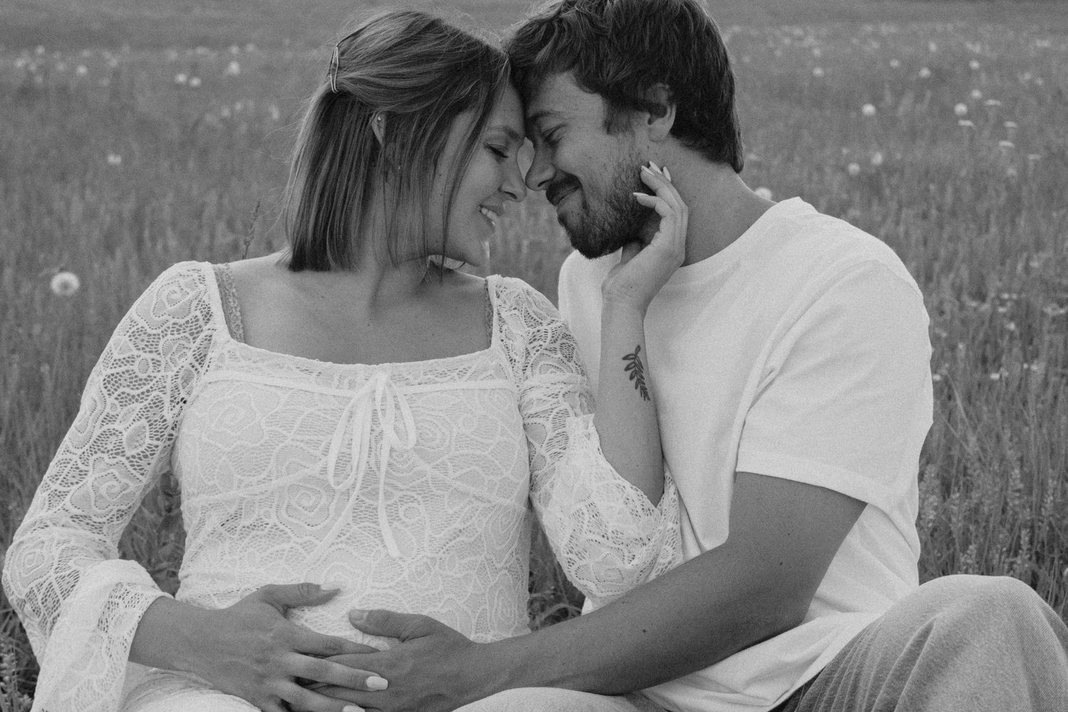 Black and white photo of a smiling pregnant couple sitting in a field, sharing a tender moment together.