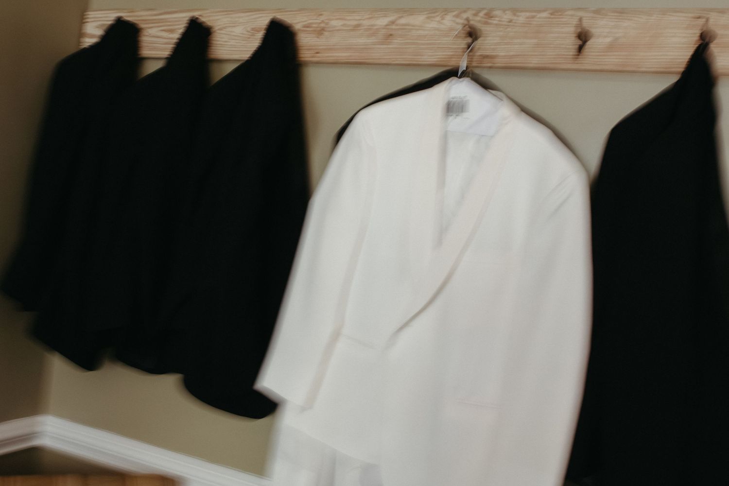 White and black formal suit jackets hanging on a rustic wooden coat rack against a neutral wall.